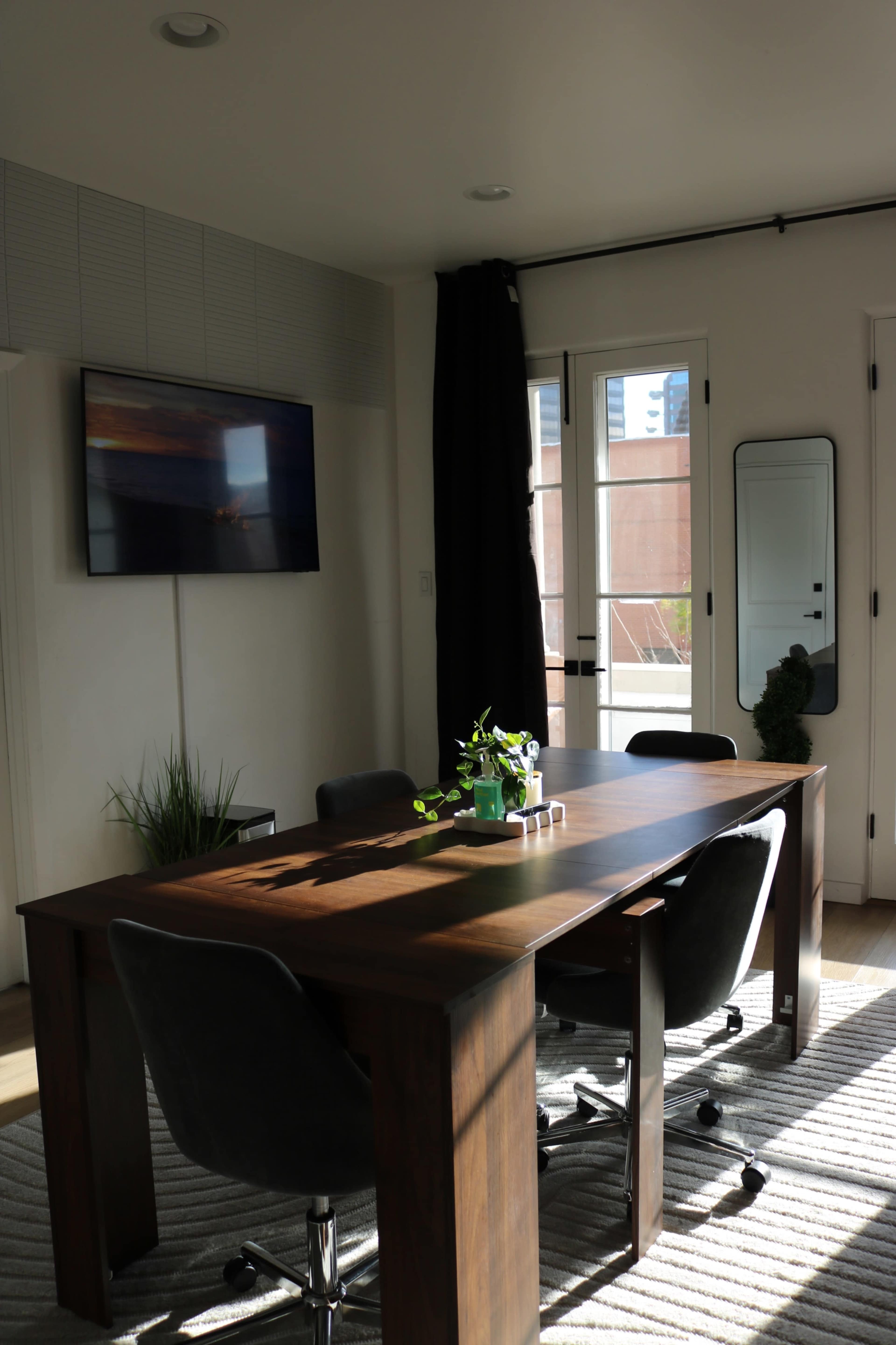 A modern meeting room features a rectangular wooden table surrounded by chairs, with a large window allowing natural light to enter.