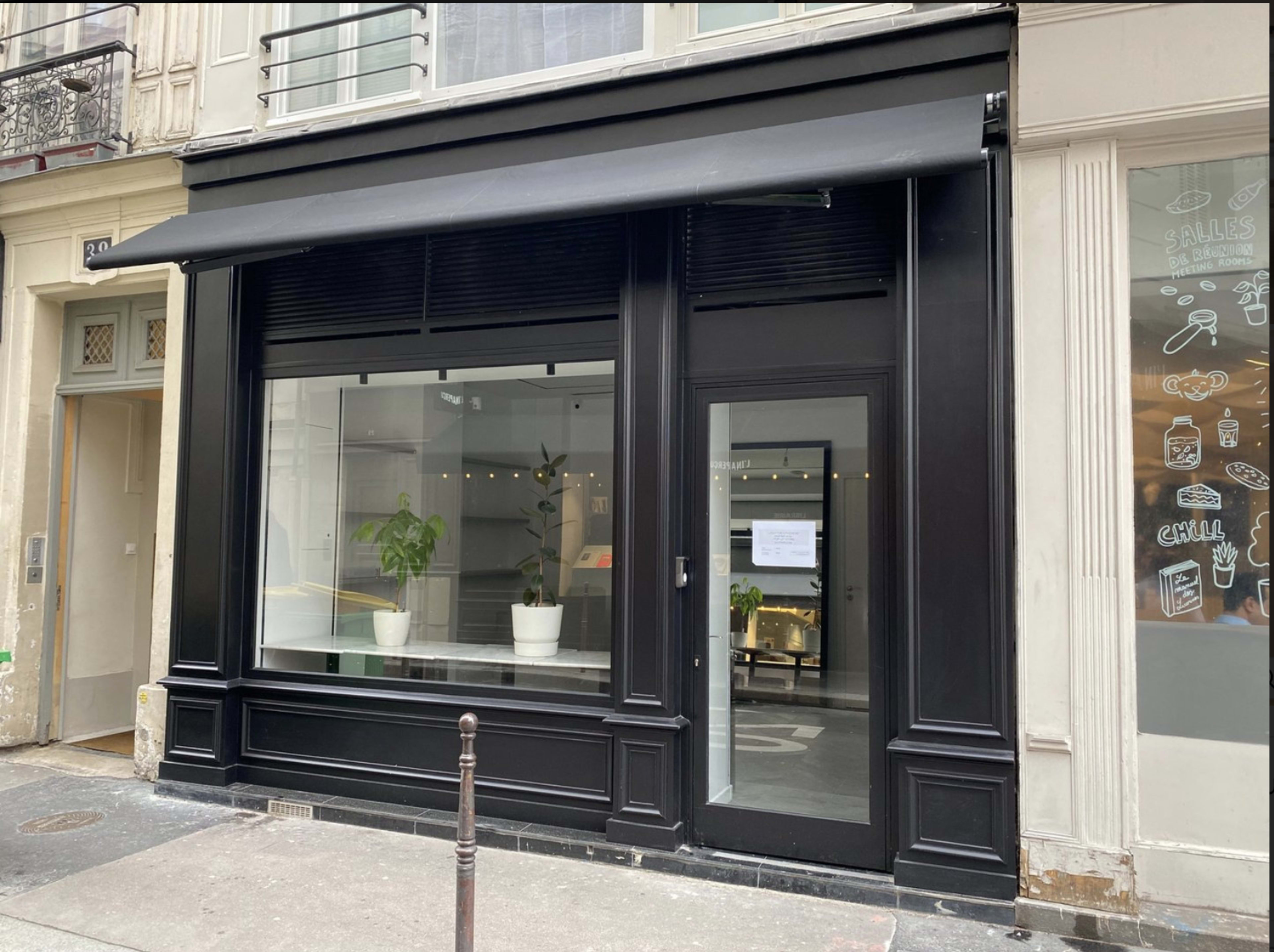 The image shows a black storefront with large glass windows and a minimalist design, located on a street with buildings on either side.