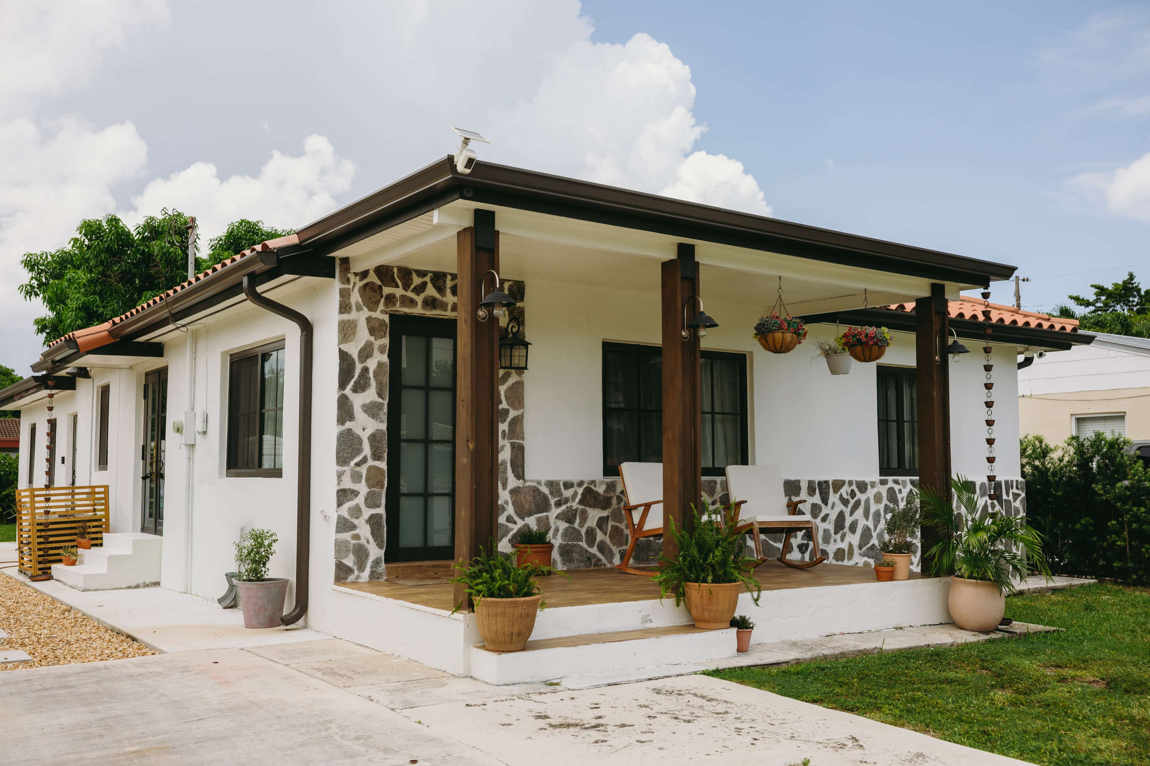 A single-story house features a stone-accented exterior, a covered porch with two chairs, and potted plants on either side.