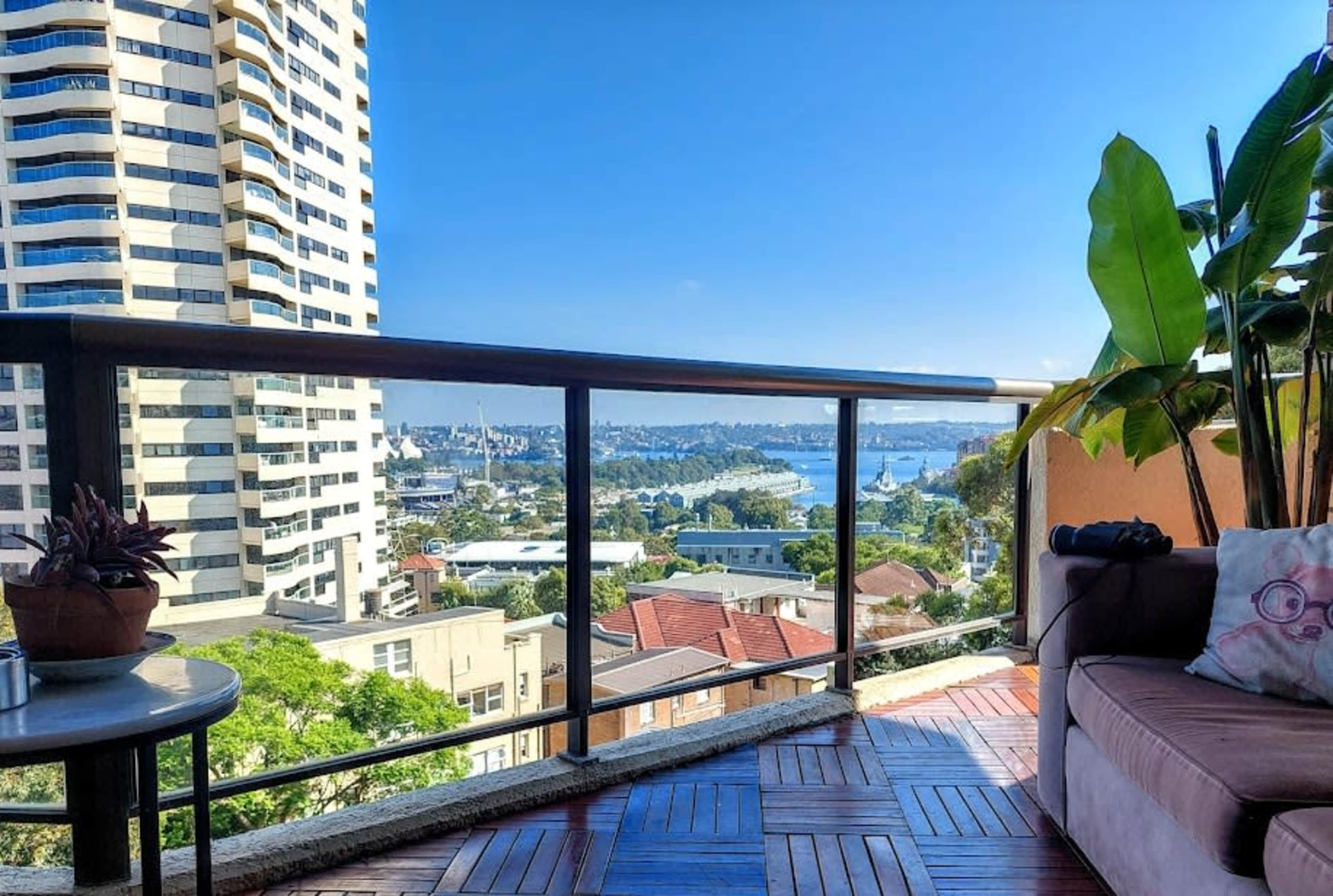 A balcony view of a city skyline featuring tall buildings, a body of water, and lush greenery.