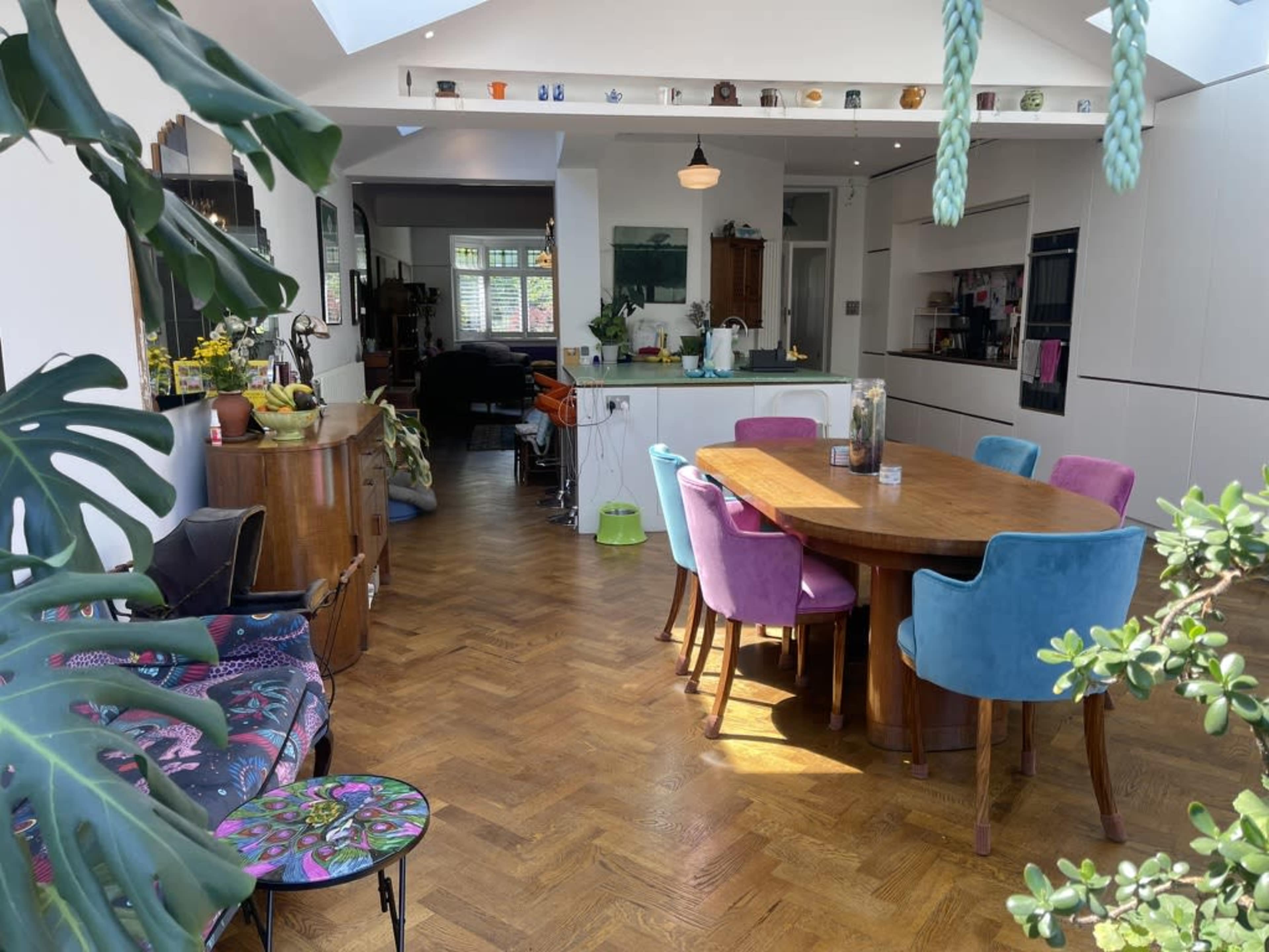The image shows a bright, open-plan kitchen and dining area featuring a wooden table surrounded by colorful chairs, with plants visible in the foreground.