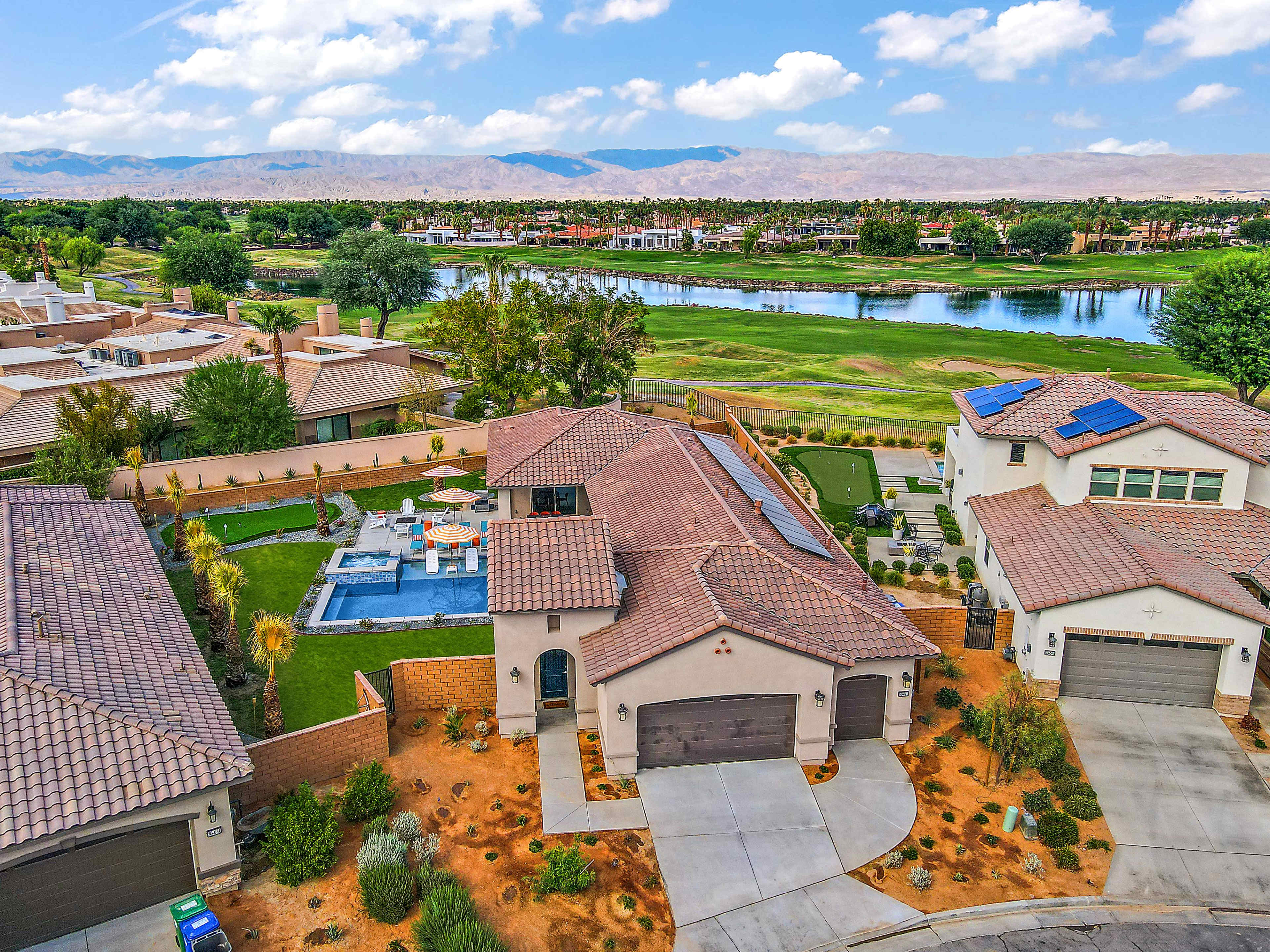 A residential neighborhood featuring several houses, a swimming pool in the backyard, and a lake with mountains in the background.