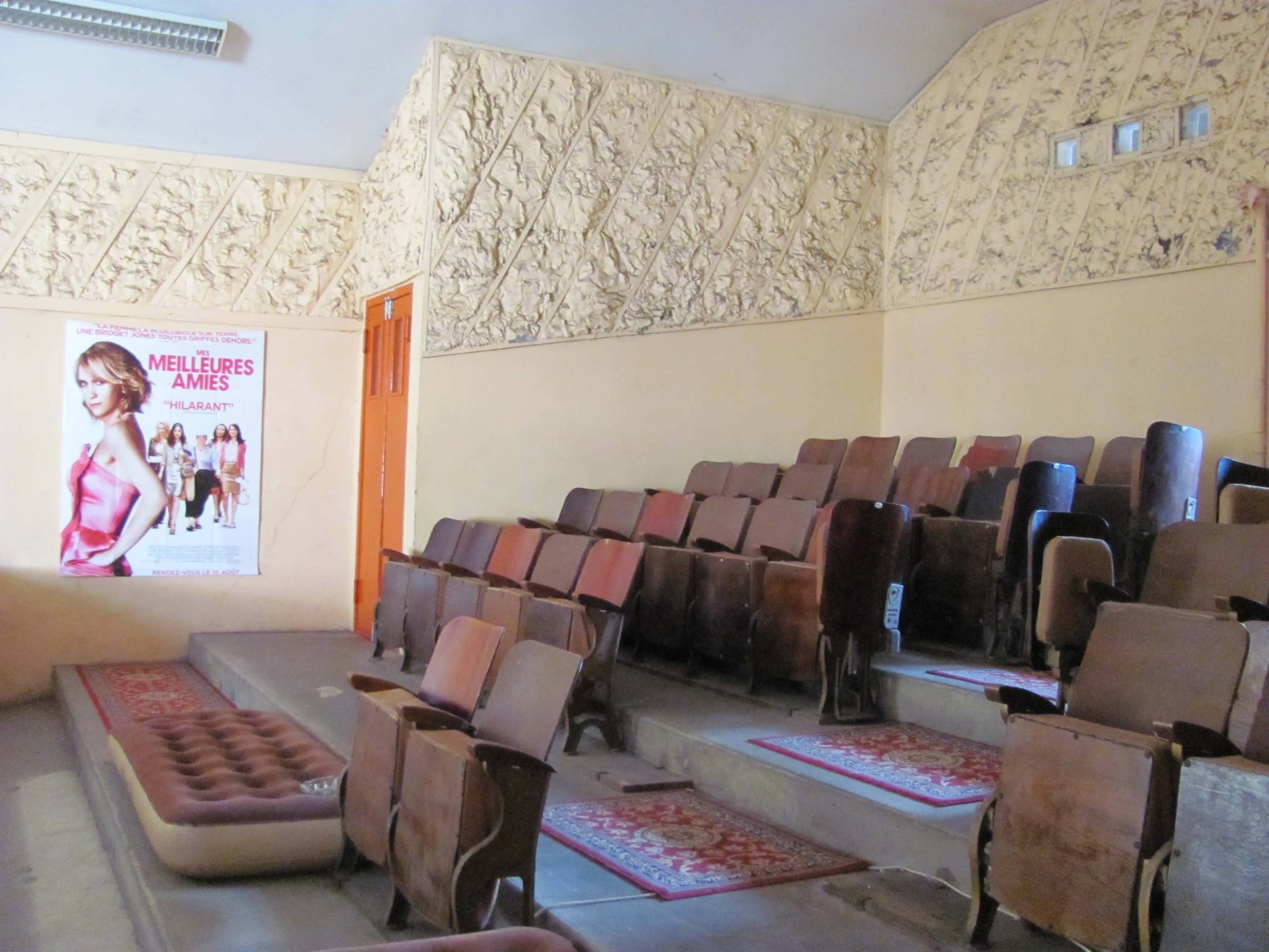 A small, empty cinema with rows of wooden seats and a decorative textured wall, along with a movie poster on one side.