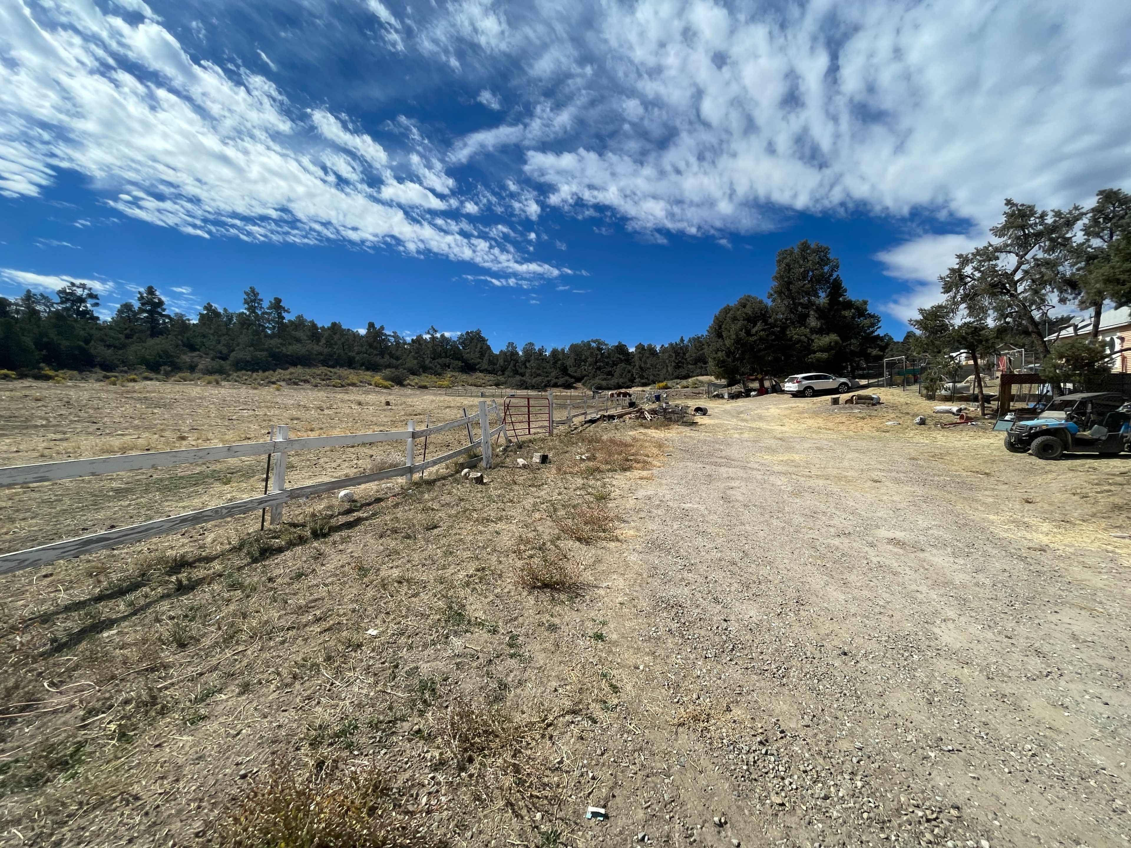 A gravel road lined with a white fence leads into a sparse, open area with scattered vehicles and trees under a partly cloudy sky.