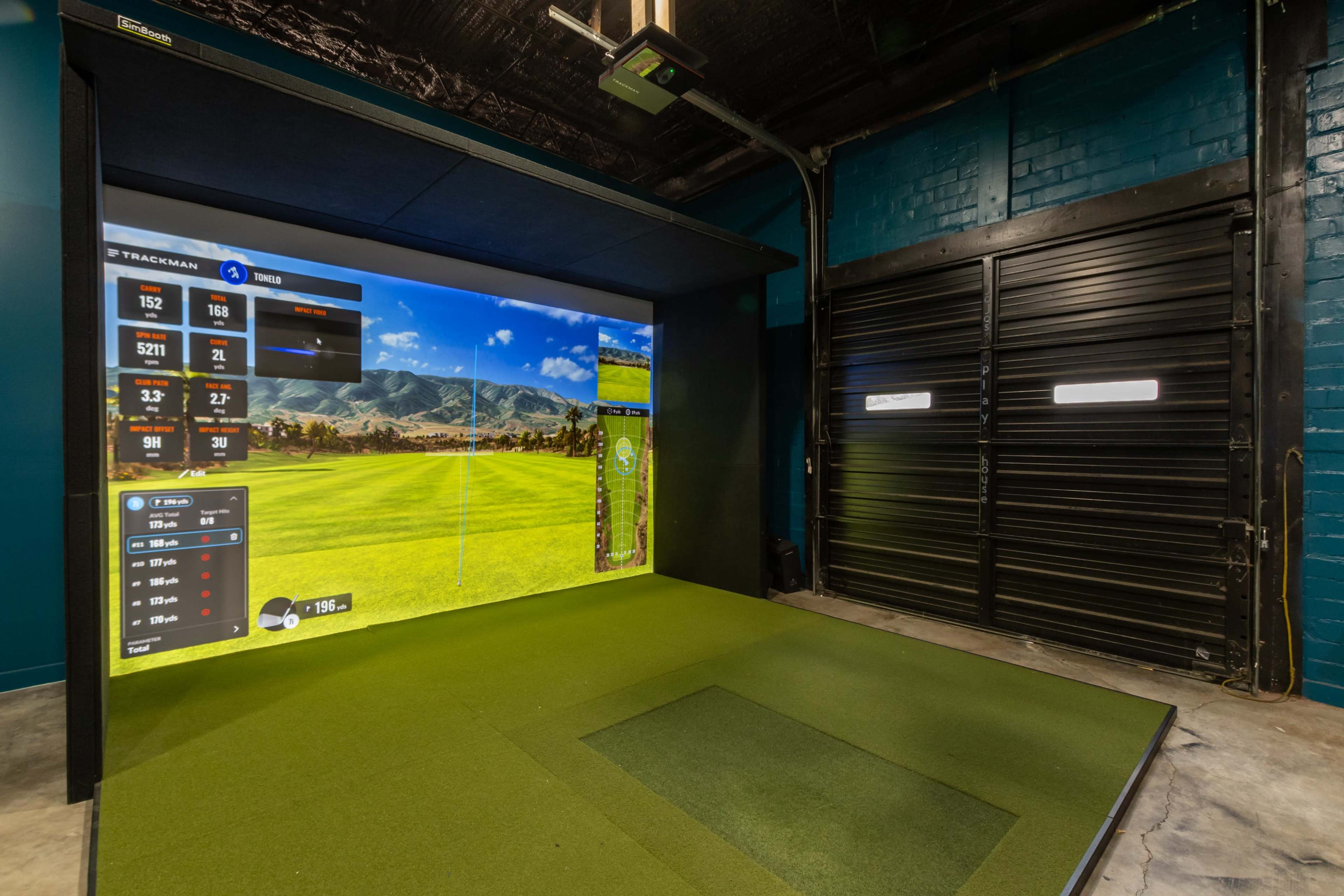 An indoor golf simulation room featuring a large screen displaying a golf course, along with a putting green area and a black garage door.