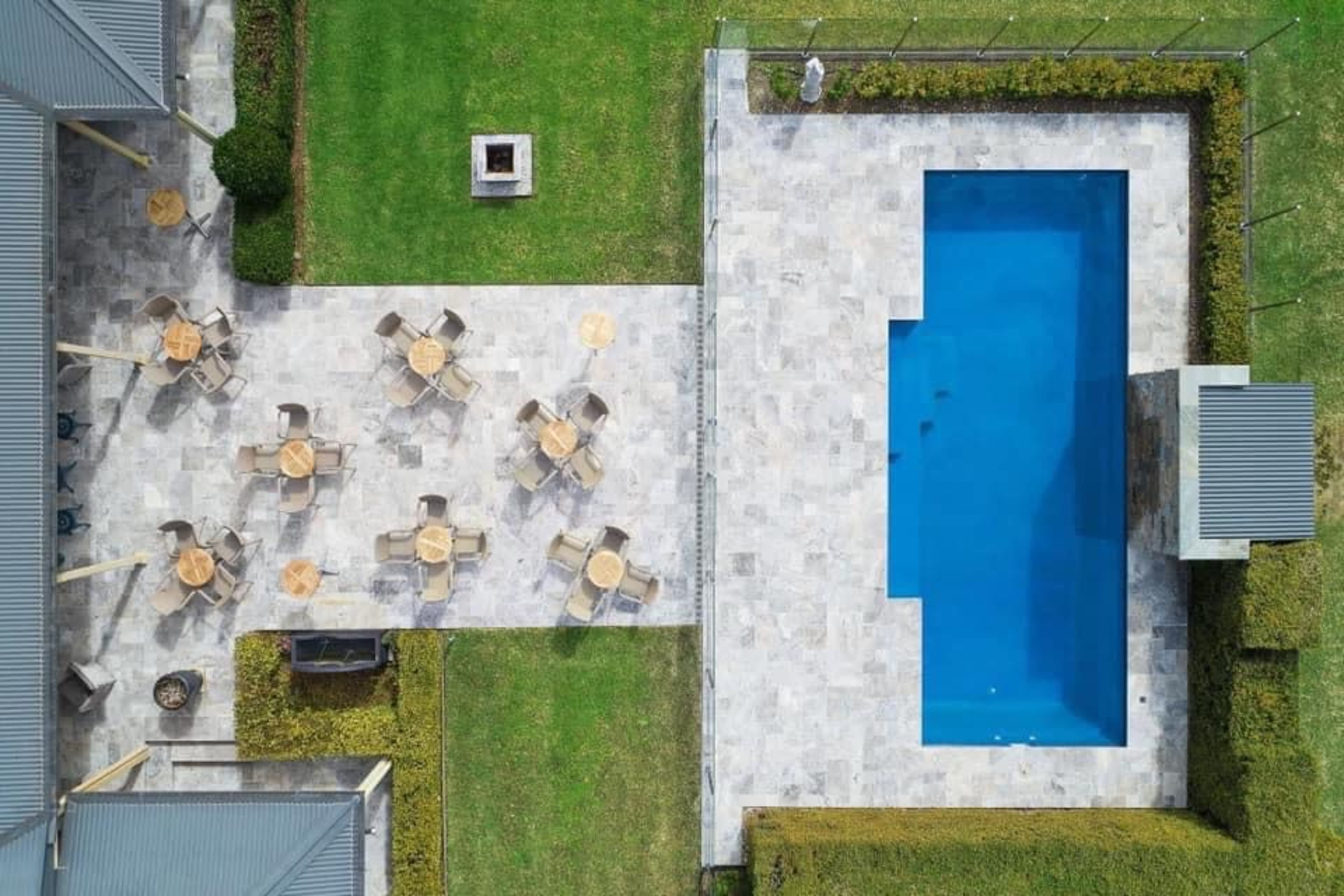 An aerial view shows a modern backyard featuring a rectangular swimming pool surrounded by a stone patio with several tables and chairs.