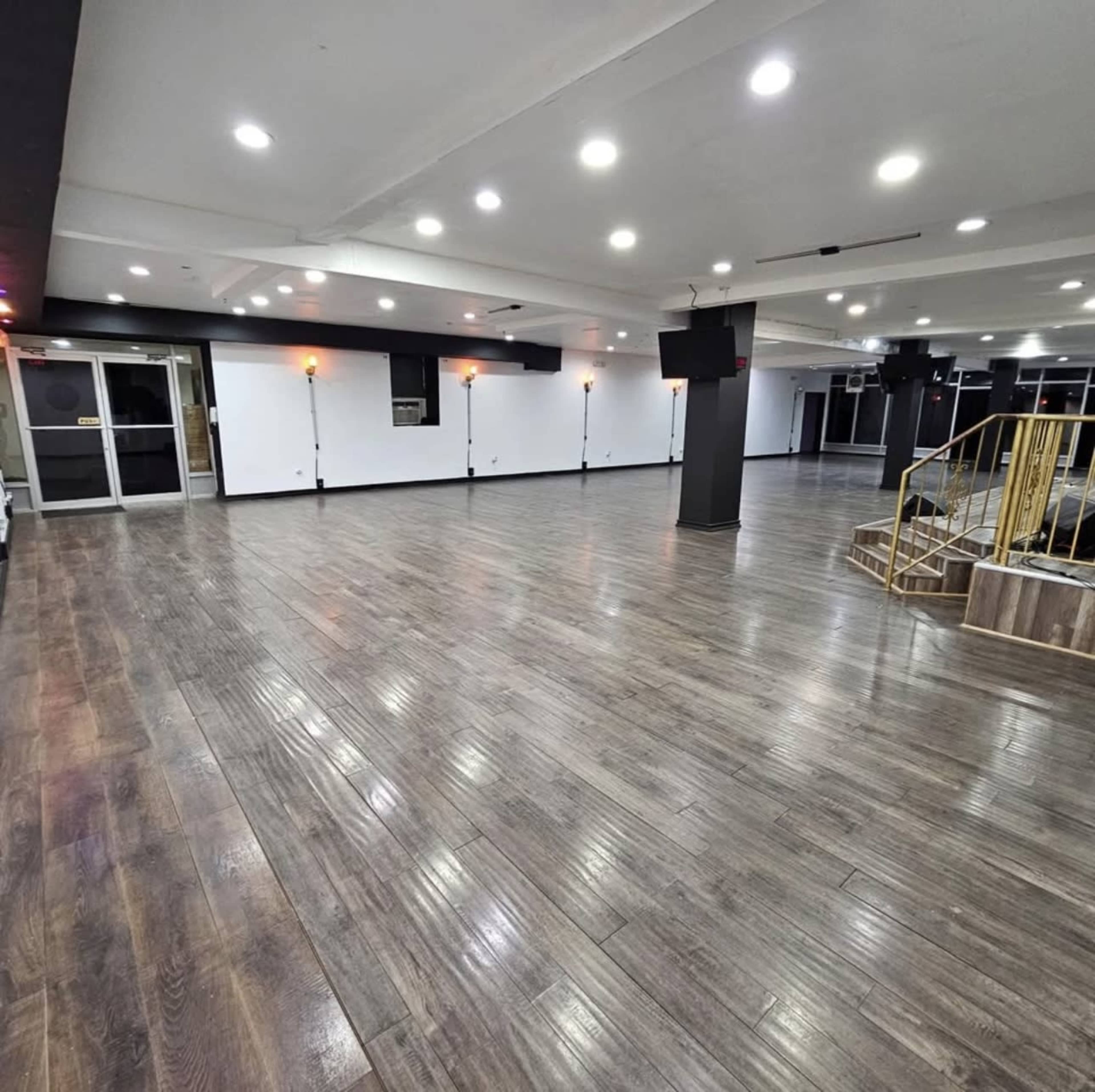 The image shows a spacious interior with polished wooden floors, bright overhead lights, and a staircase in the corner.