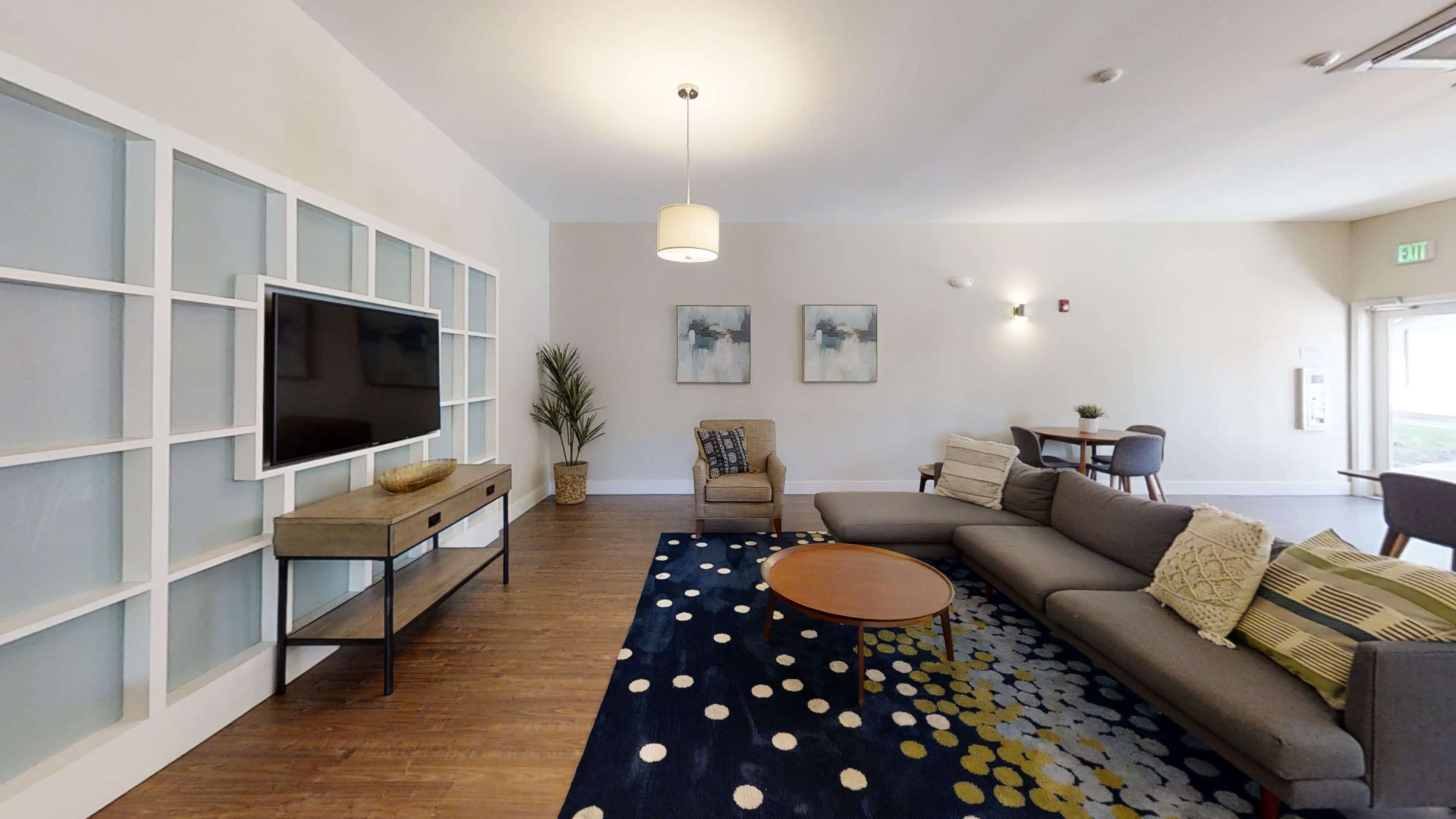 A living area with a large sectional sofa, a coffee table, a television mounted on the wall, and a dining area in the background.