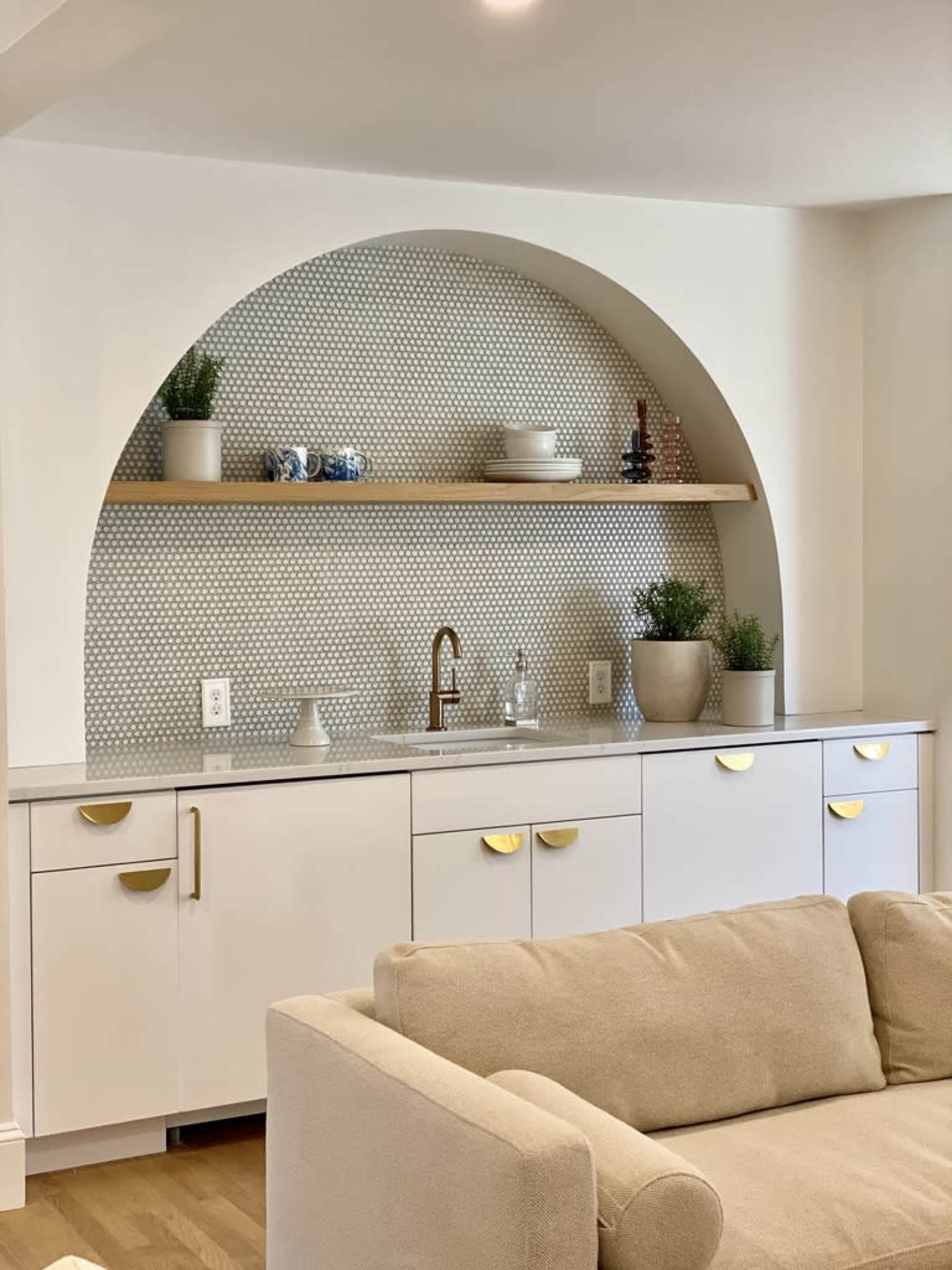 The image shows a modern kitchen area with a white cabinet and a patterned tile backsplash featuring an arch shelf displaying decorative items and potted plants.