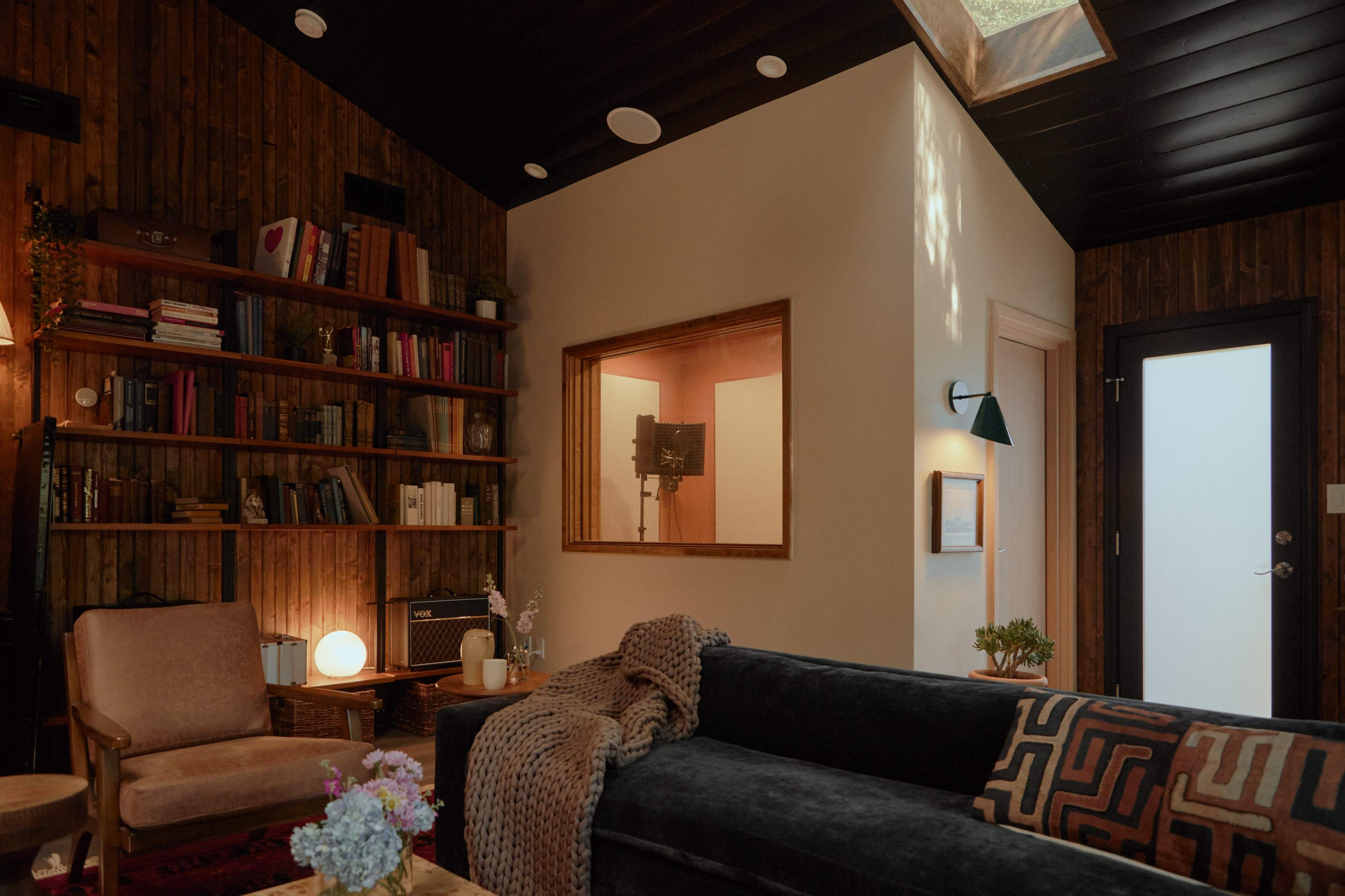 A cozy living room with wooden walls, bookshelves filled with books, a sofa, and a large window illuminating the space.