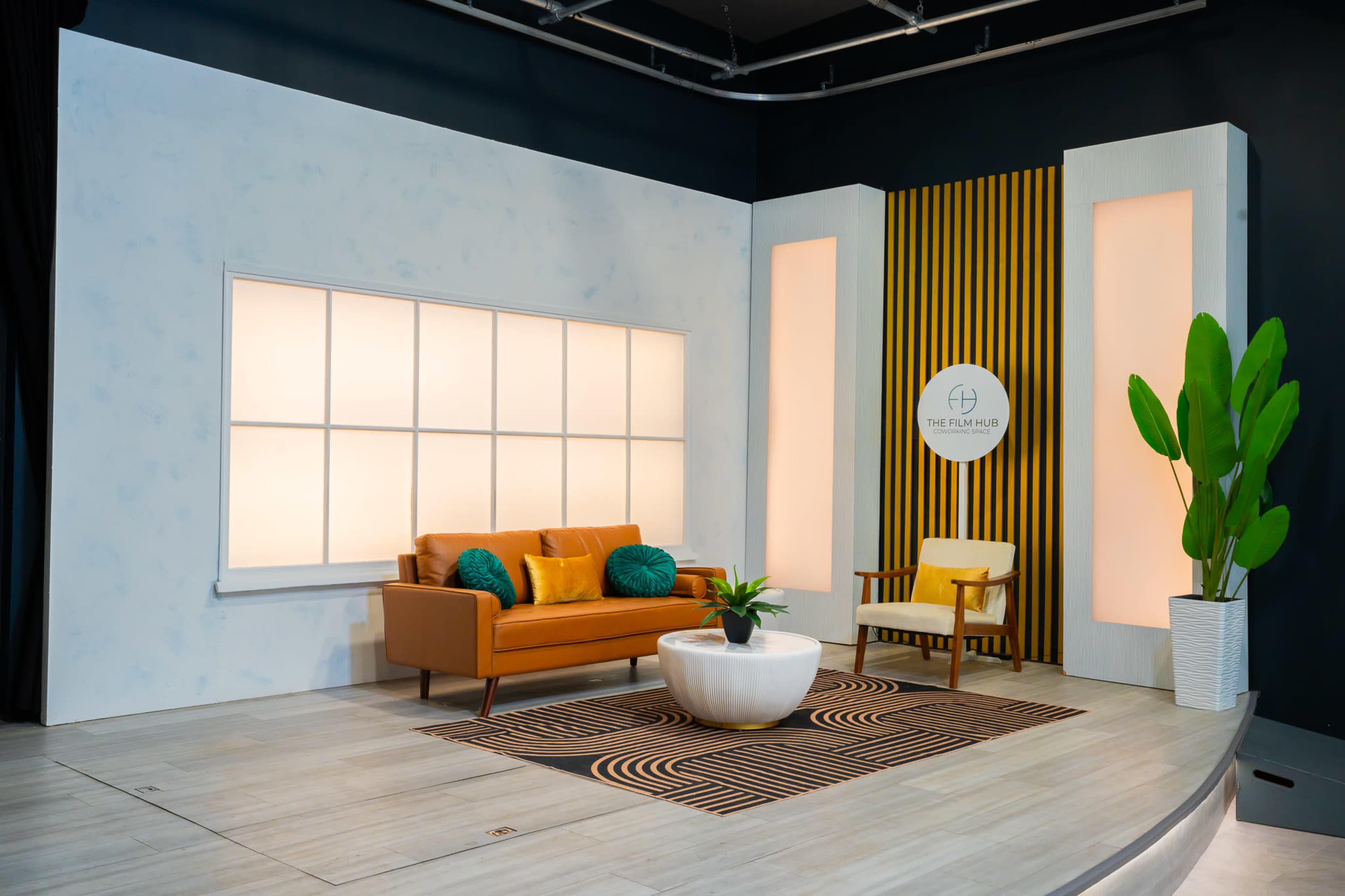 The image shows a modern living room setting with a brown sofa, a round white coffee table, a plant, and a striped backdrop featuring a circular logo.