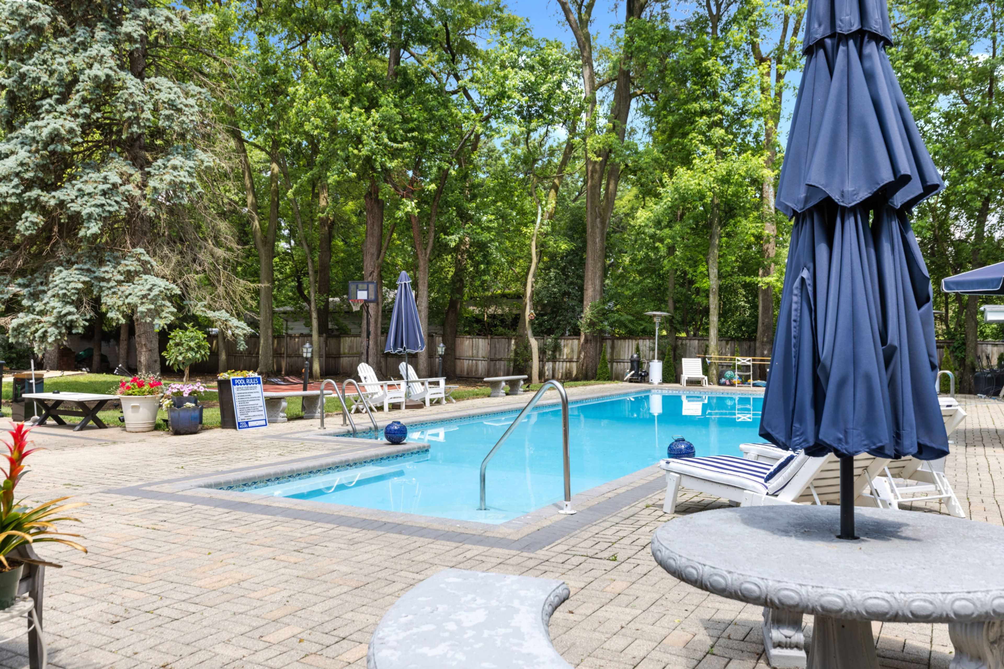 The image shows a clear swimming pool surrounded by lounge chairs and umbrellas, set in a landscaped area with trees.