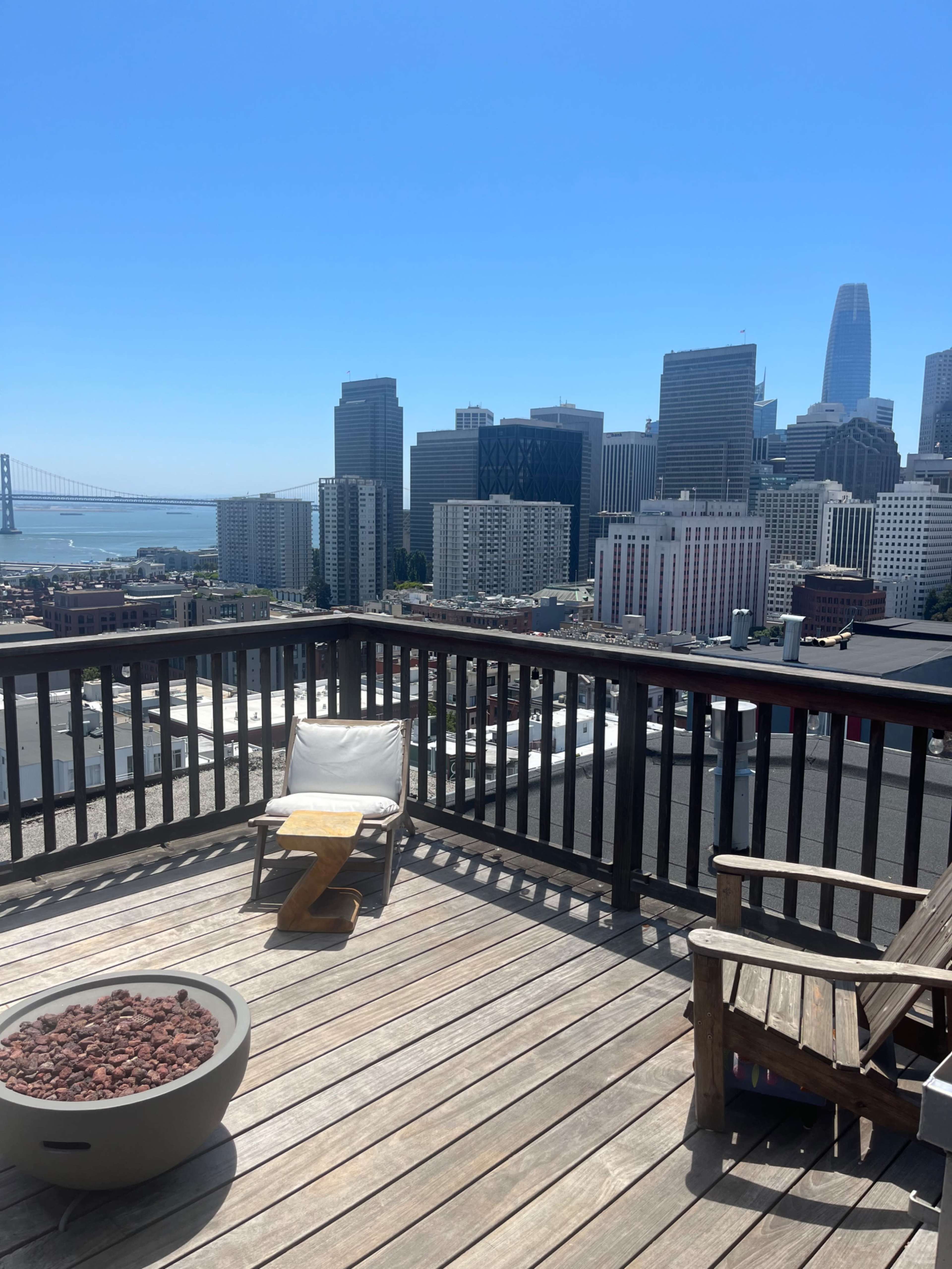 A deck featuring a fire pit and a lounge chair overlooks a city skyline with skyscrapers and a bridge in the distance.
