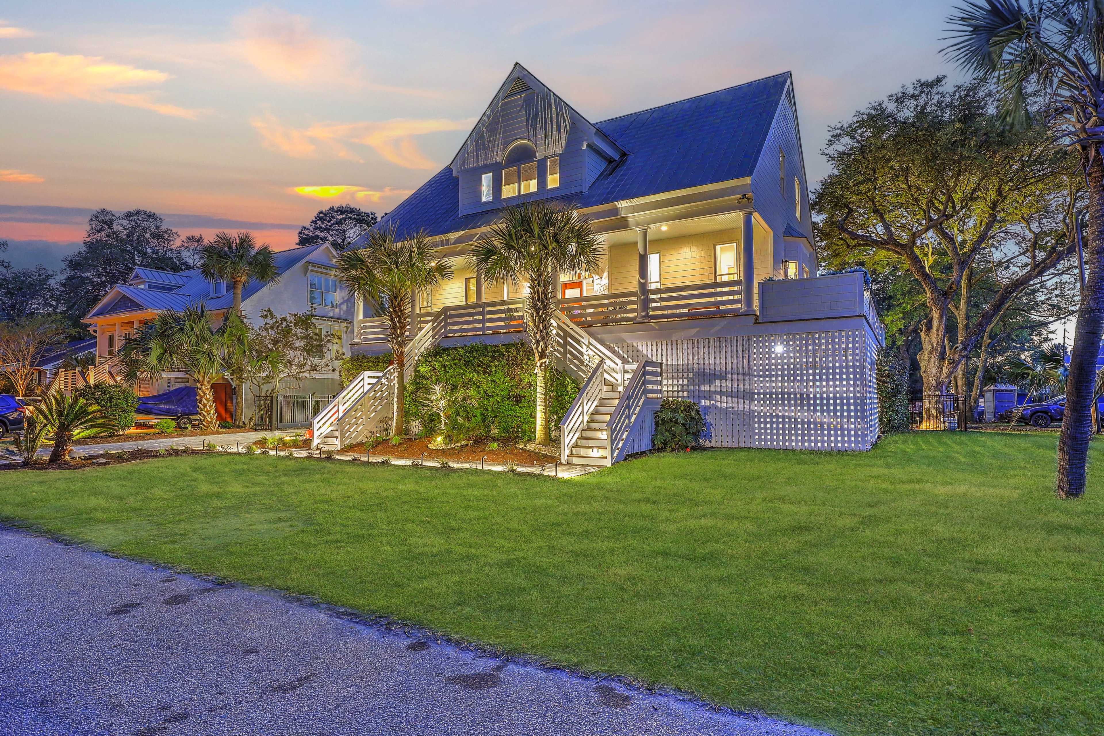A large, two-story house with a steep roof and a wooden deck is surrounded by palm trees and lush green grass under a colorful sunset sky.