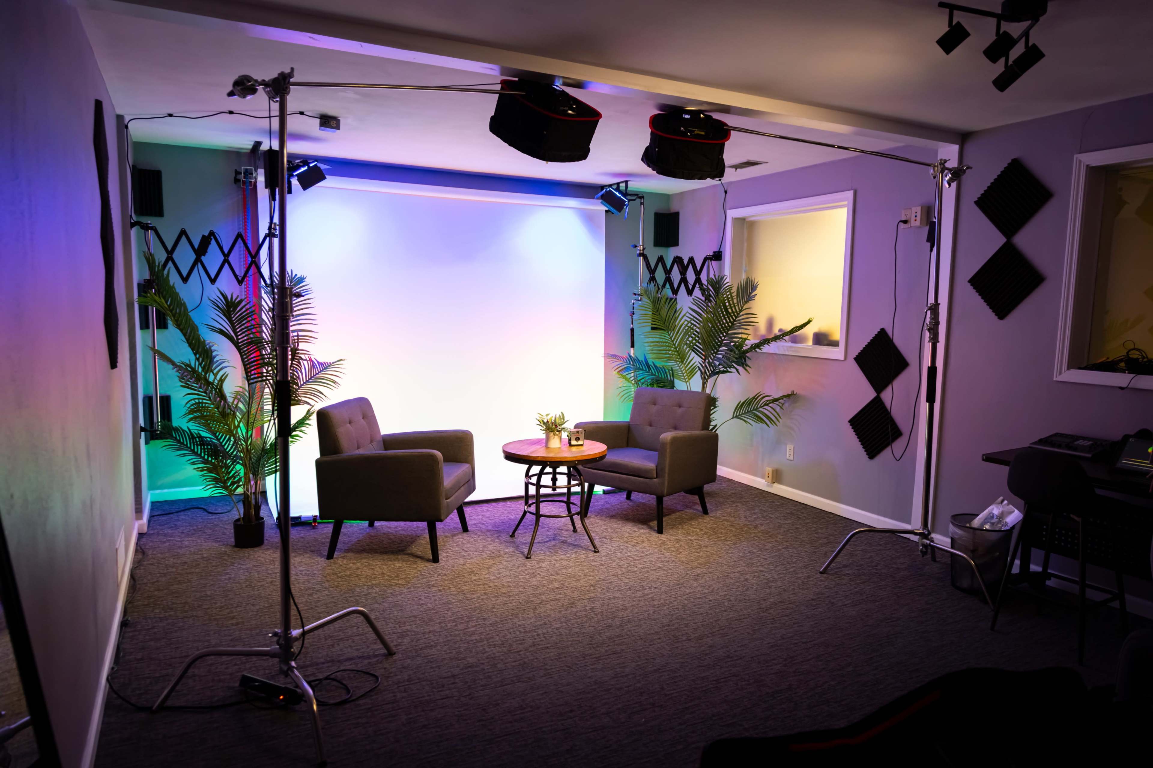 A studio setting featuring two grey chairs, a small table with a plant, and colorful lighting in the background.
