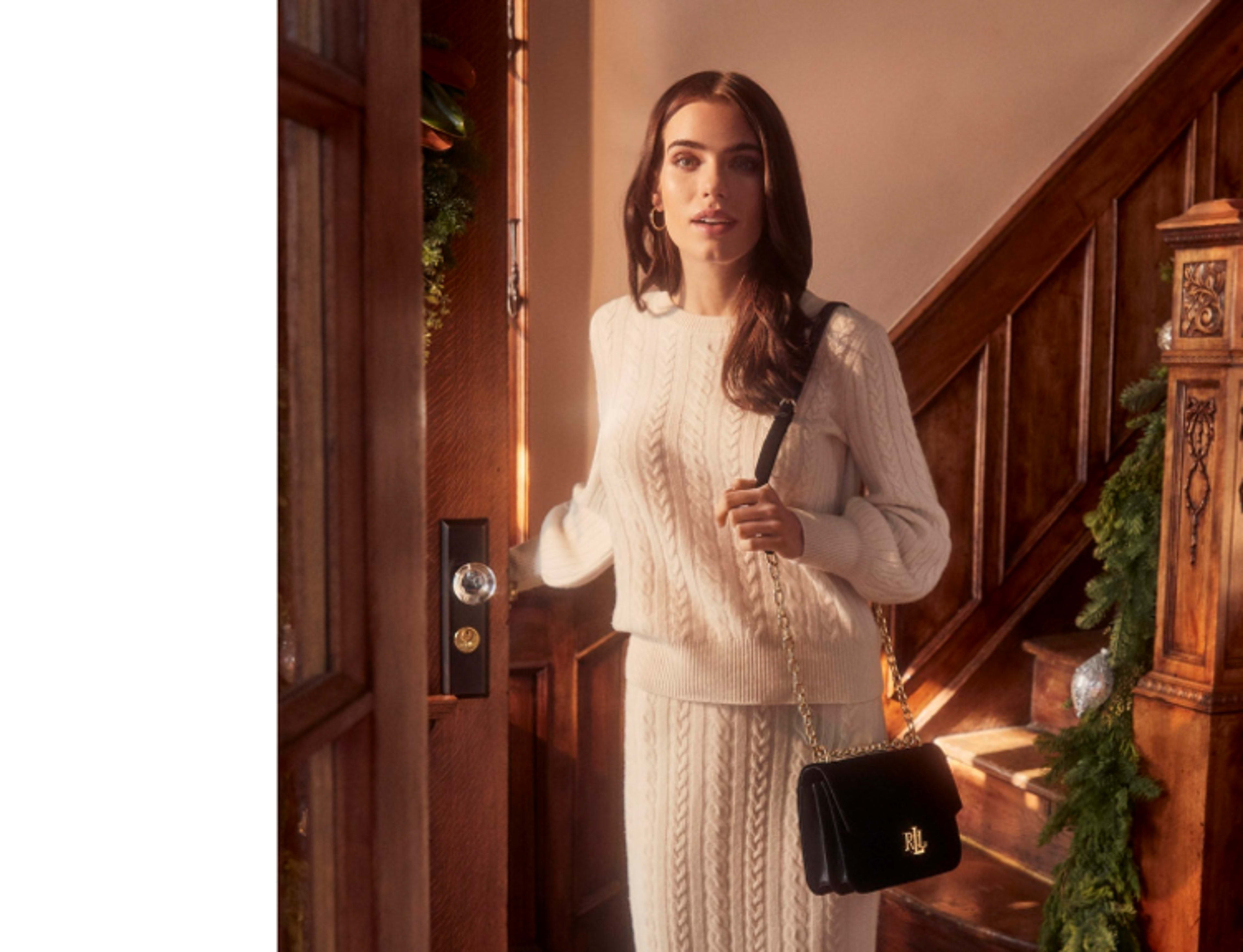 A woman wearing a cream-colored knitted sweater and skirt stands by an open door, holding a black handbag, with a staircase and holiday decorations visible in the background.
