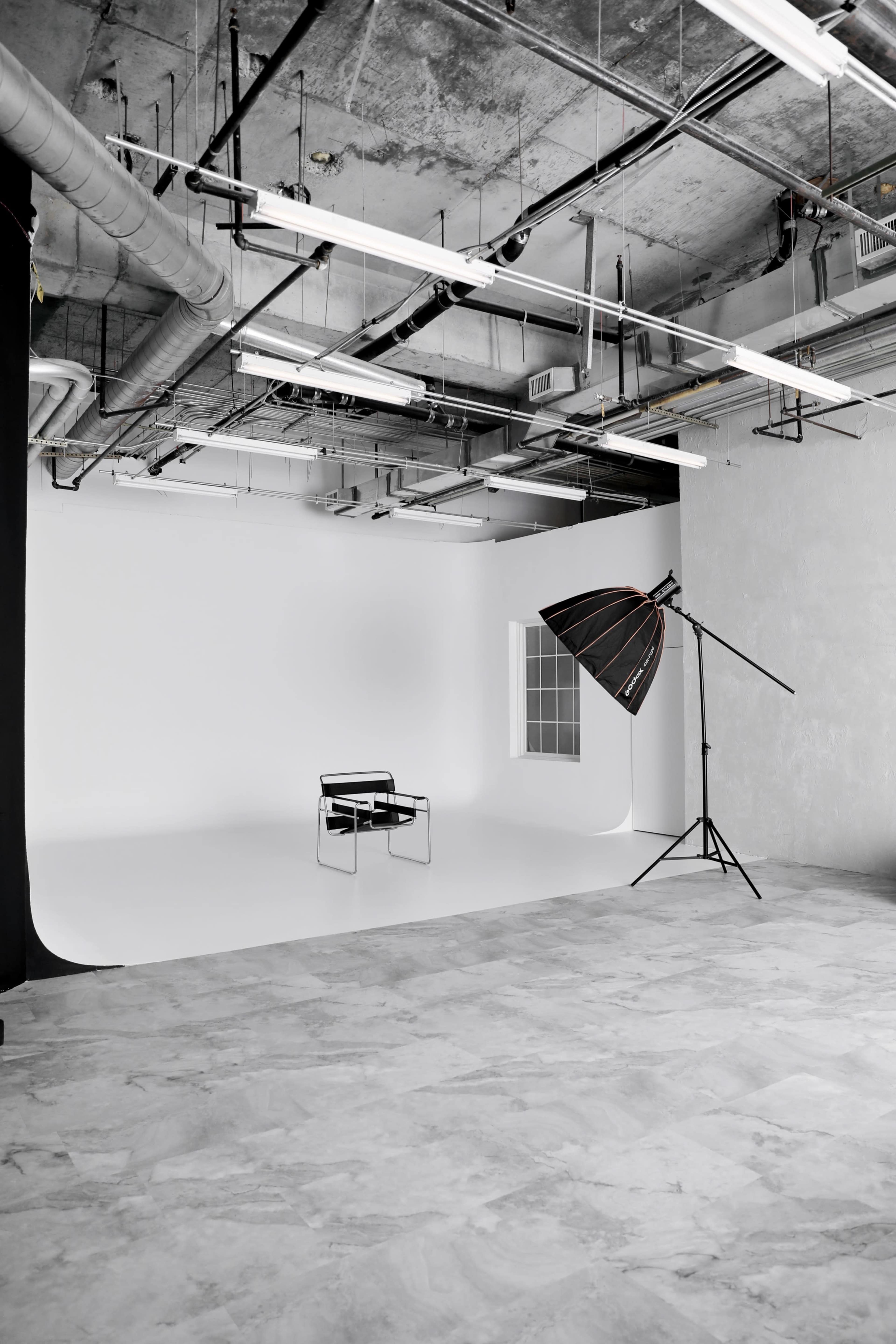 The image shows a minimalist photography studio with a black chair, a large softbox light on a stand, and a blank wall in the background.