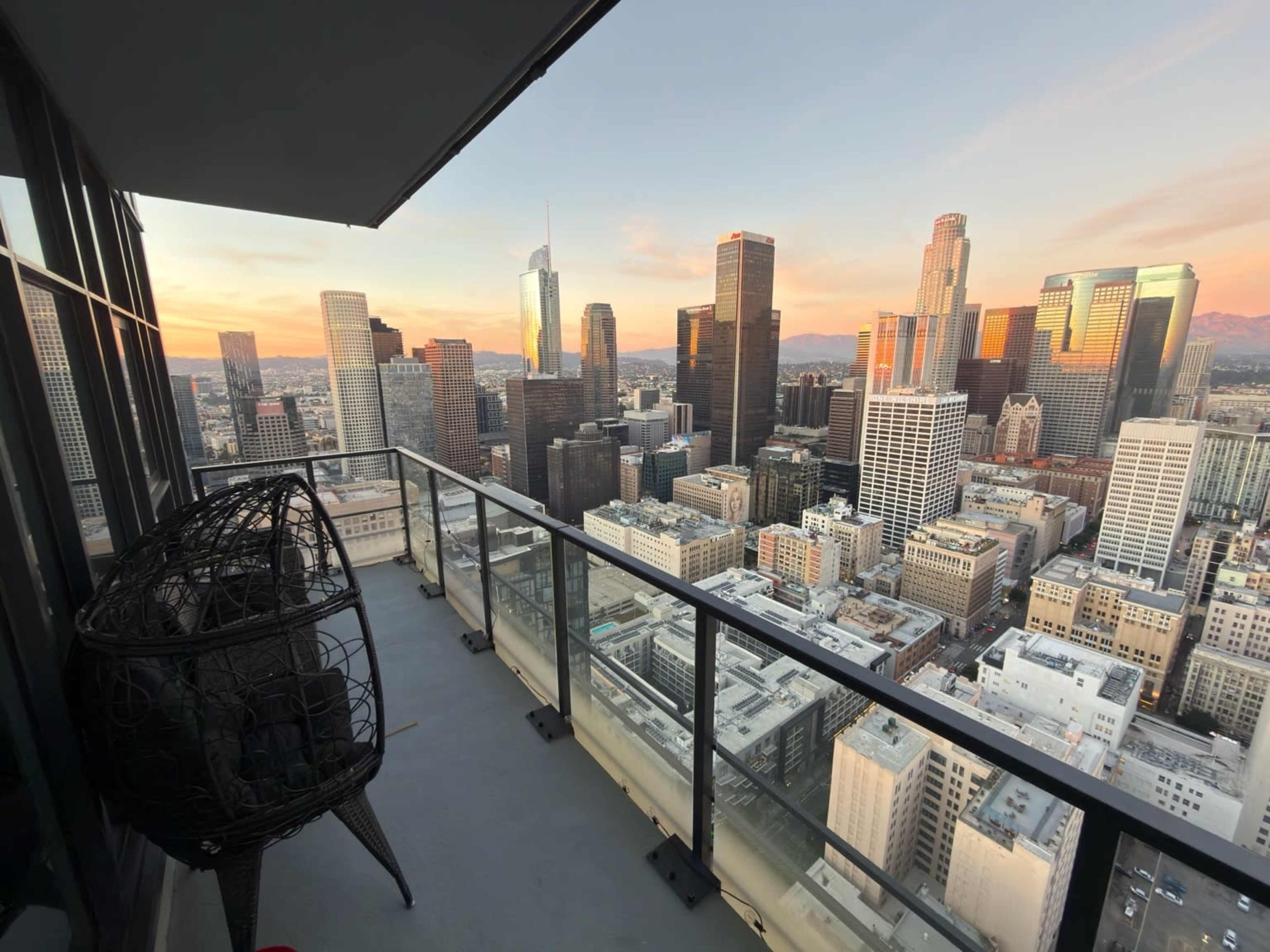 The image shows a high-rise balcony in downtown Los Angeles, overlooking a skyline filled with skyscrapers during sunset.
