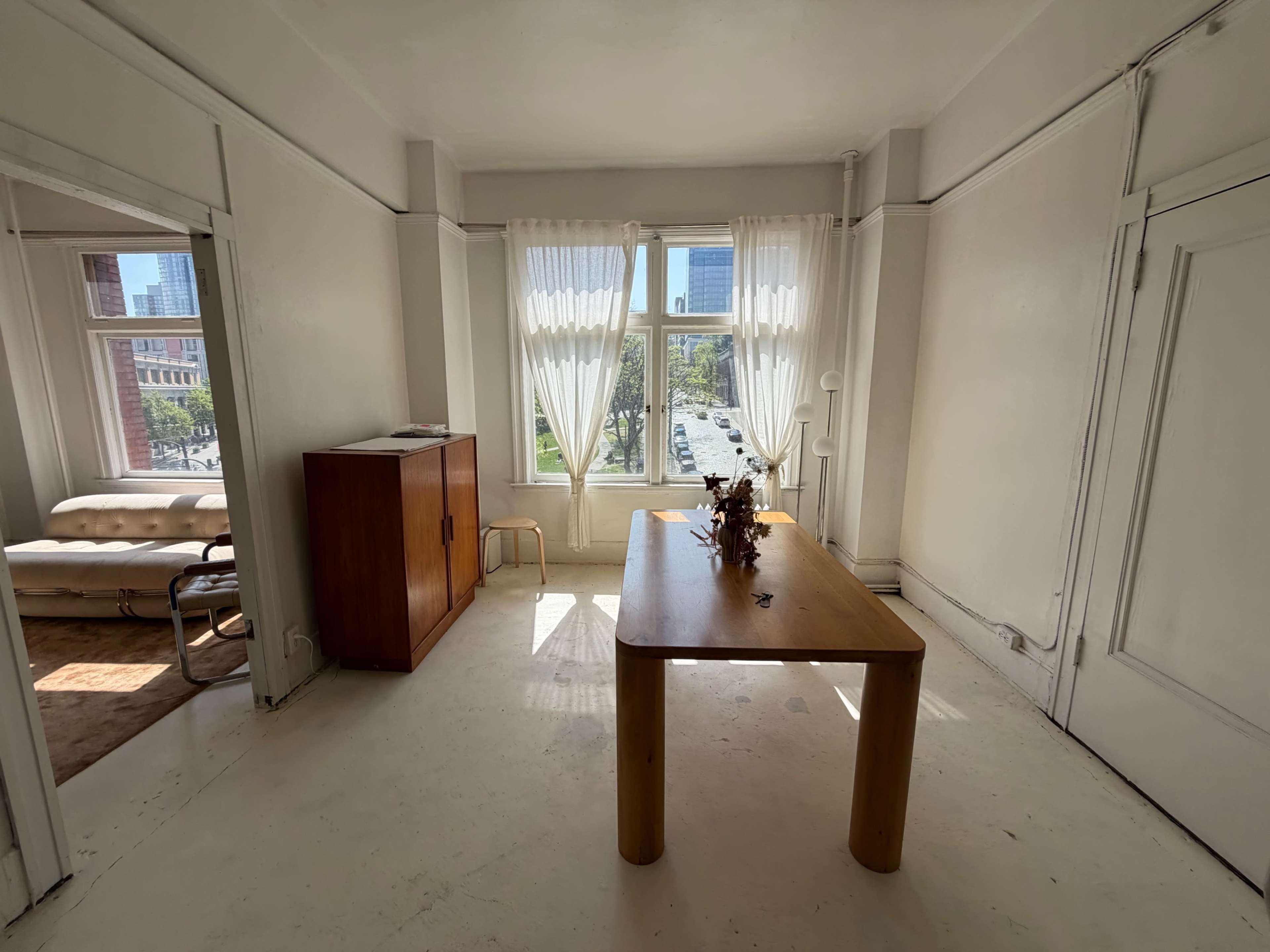 The image shows a sunlit room with a wooden table and a cabinet, featuring large windows draped with sheer curtains.