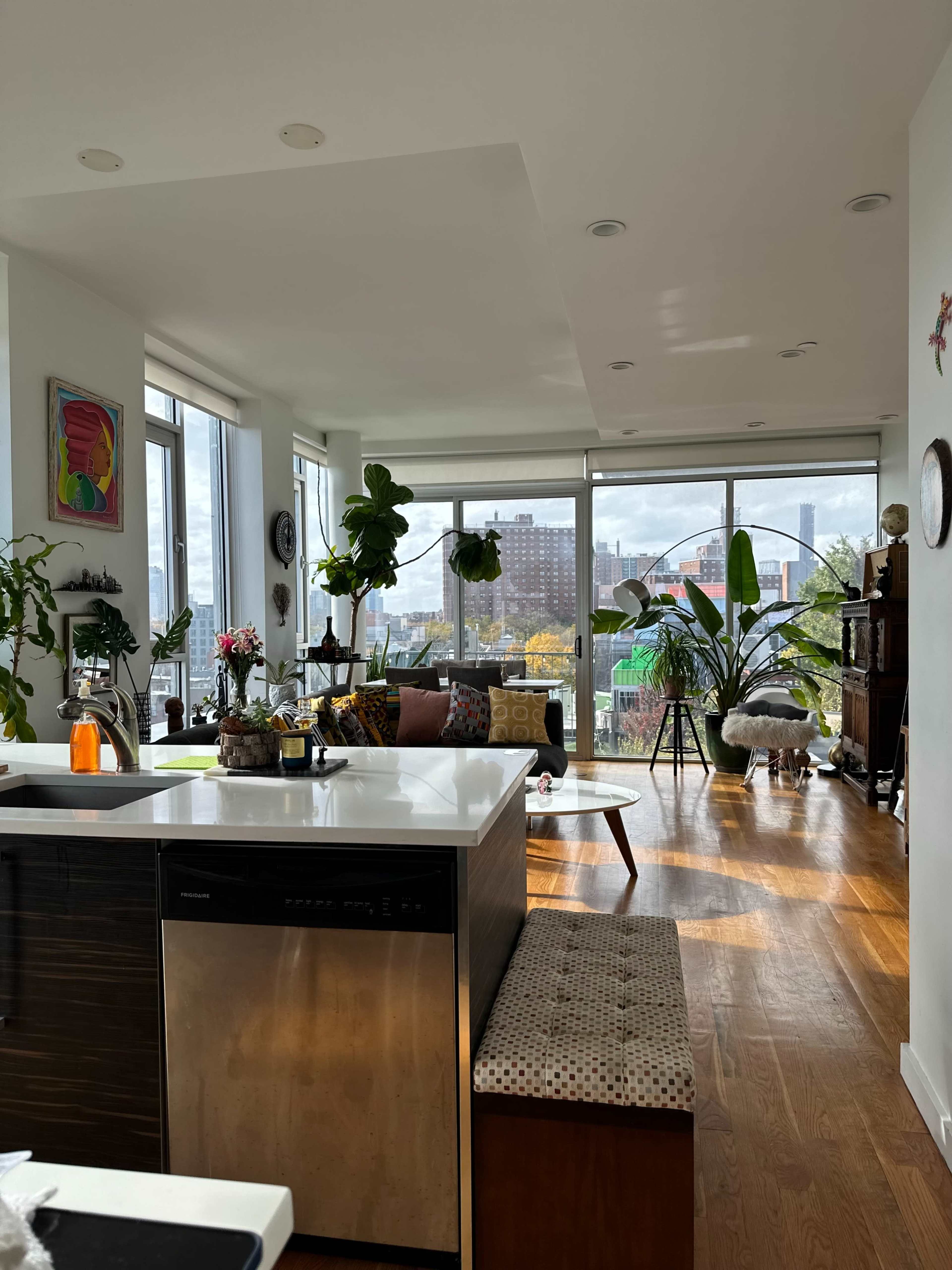 The image shows a modern open-concept kitchen and living area with large windows, plants, and a view of the city skyline.