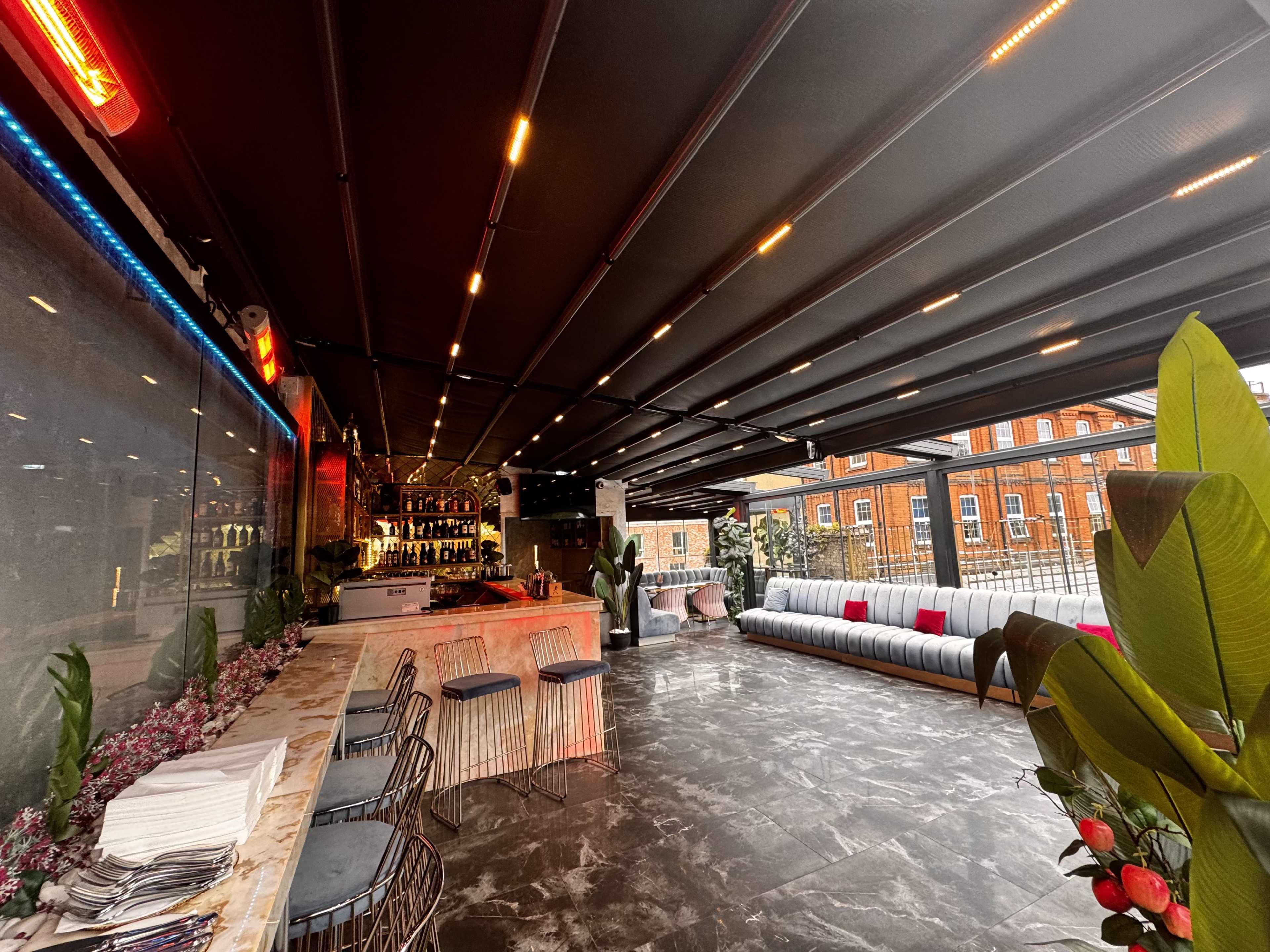 The image shows a modern rooftop bar with a sleek design, featuring a bar counter, seating area, and plants, under a retractable ceiling.