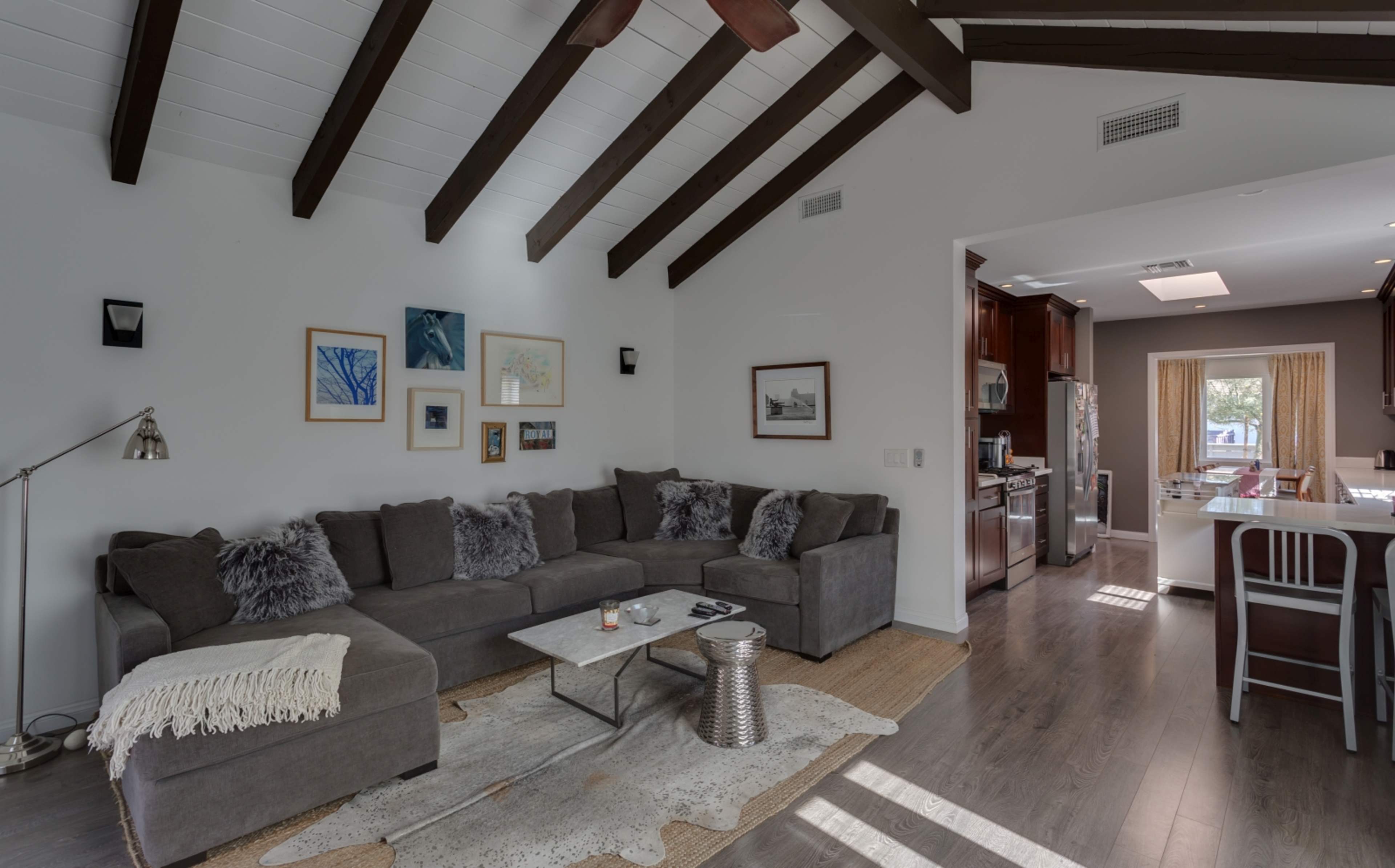 A modern living room features a sectional sofa, decorative art on the walls, and a view into a kitchen with wooden cabinets.