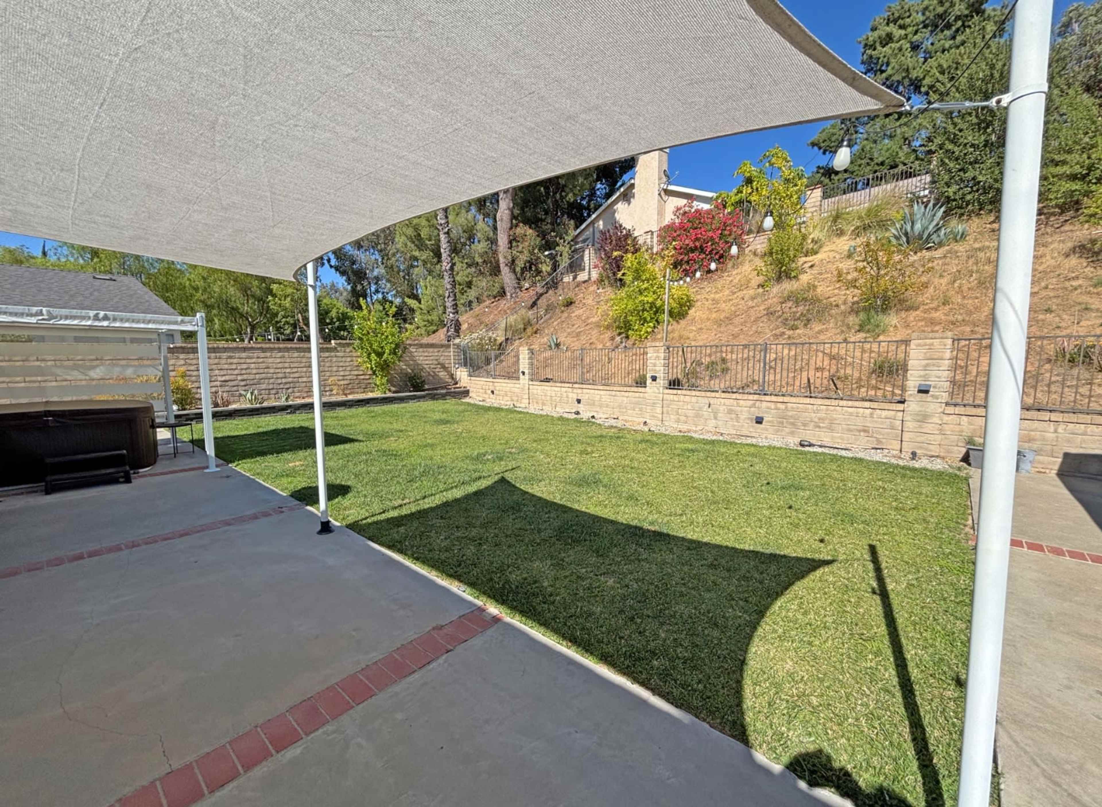 A shaded outdoor area features a grassy lawn bordered by a stone wall and plants, with a patio in the foreground.