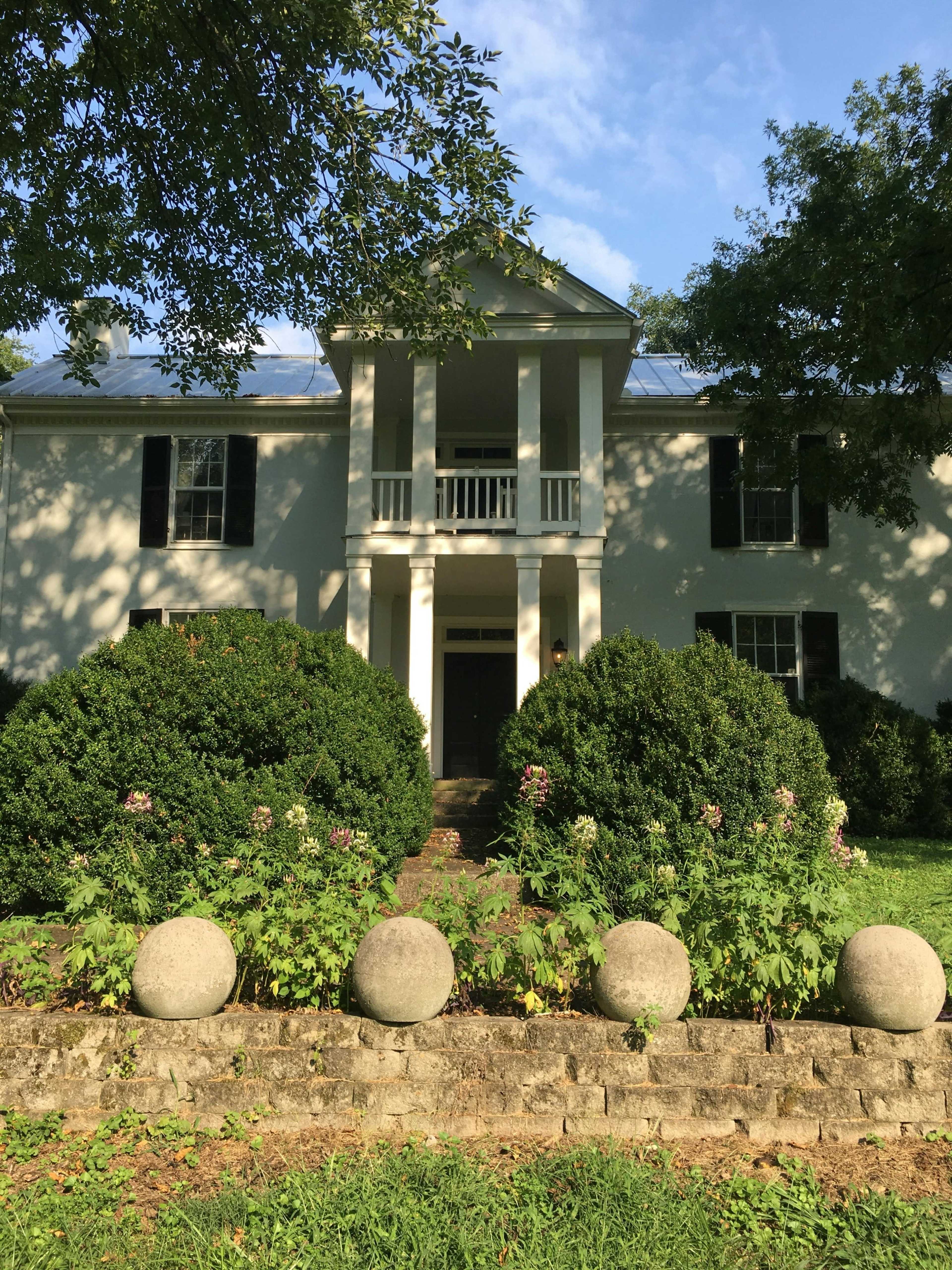 The image shows a large two-story house with a columned entrance and trees in front, surrounded by well-maintained landscaping and a stone wall with round stone accents.