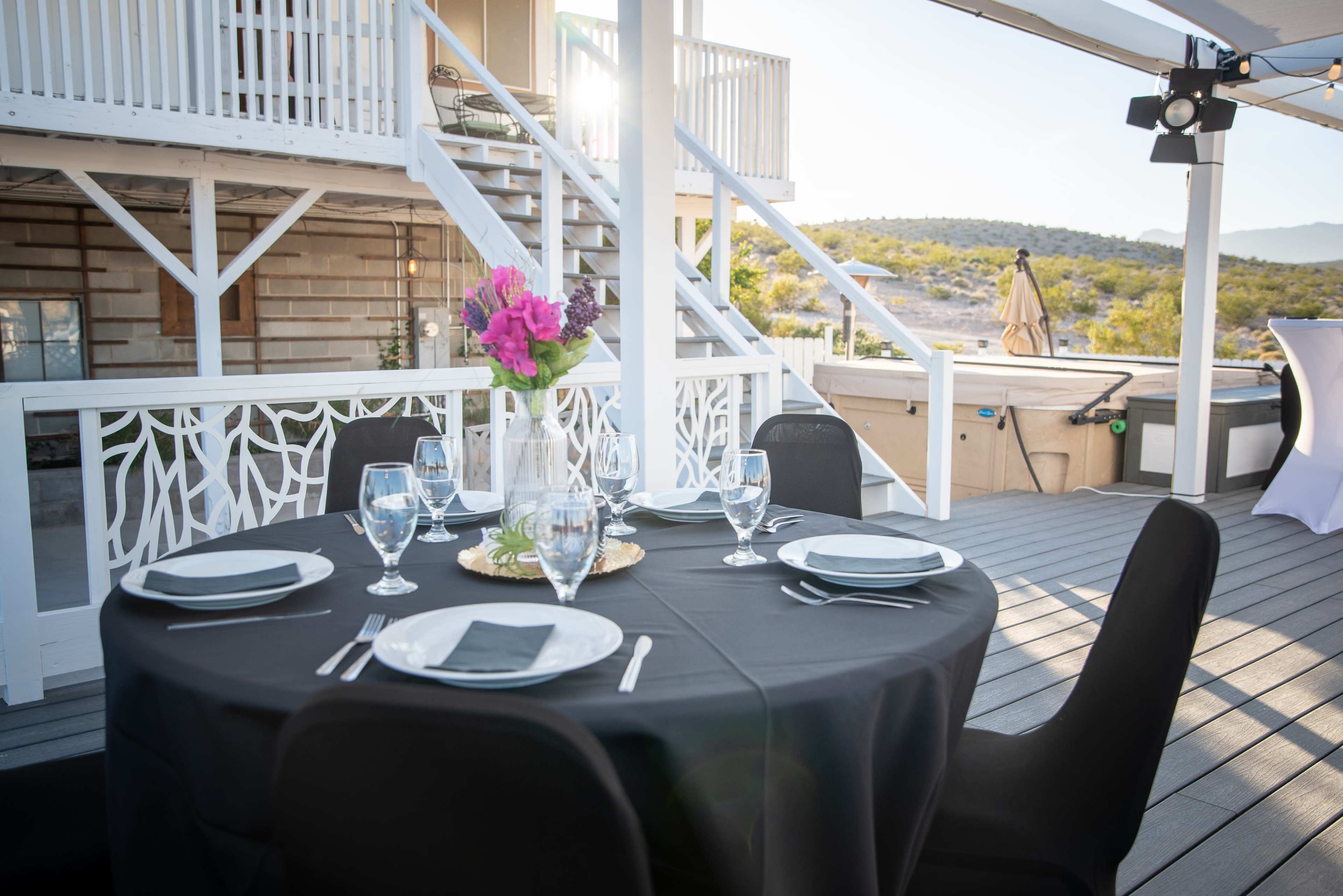 A round table is set with black linens, white plates, and glassware, accompanied by a floral centerpiece on a deck with a view of a hillside.