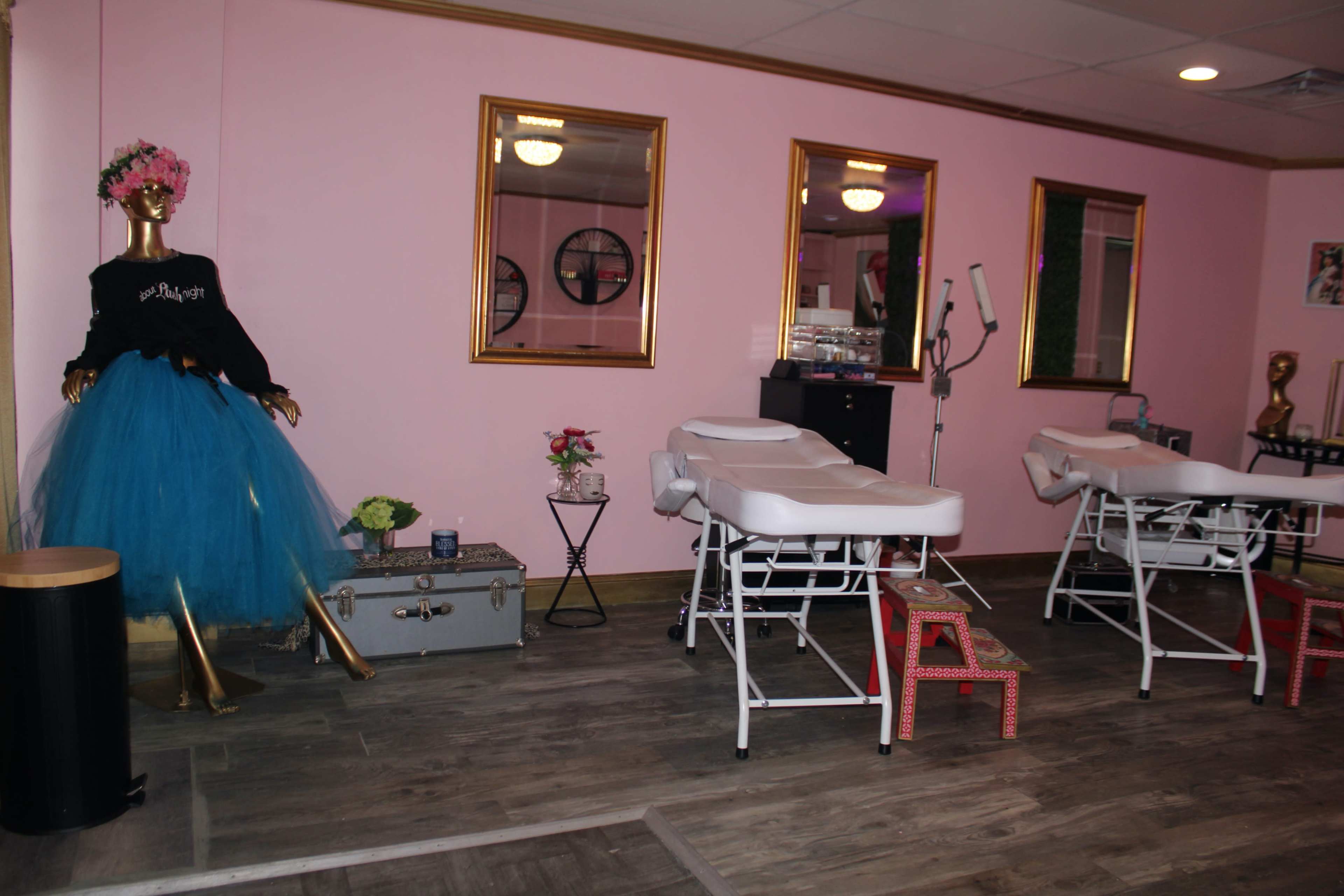 A beauty salon interior with pink walls, two treatment tables, a decorative mannequin wearing a tutu, and gold-framed mirrors.