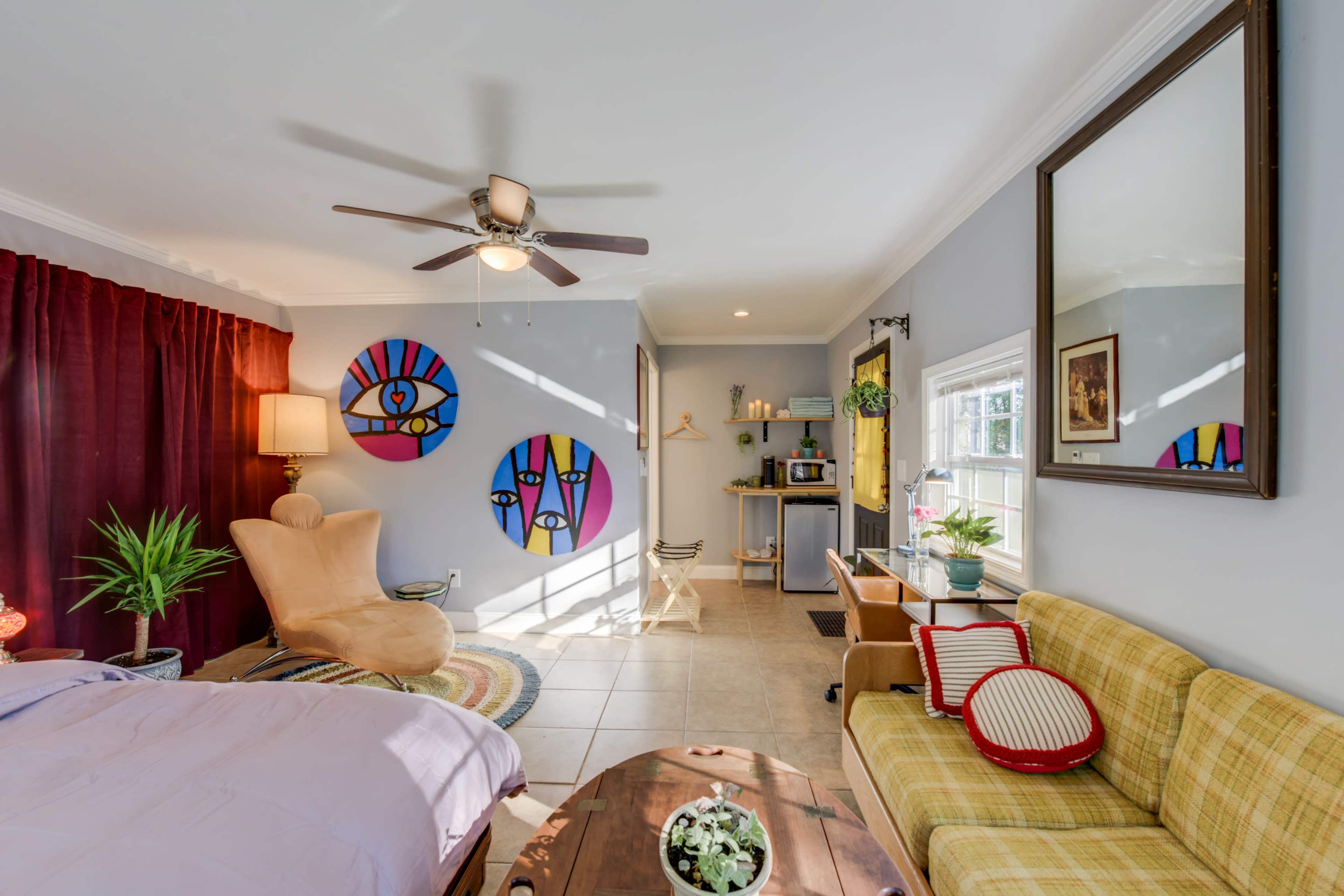 A brightly lit room features a sofa, a chair, and colorful artwork on the walls, with a kitchenette at one end and a window allowing natural light.