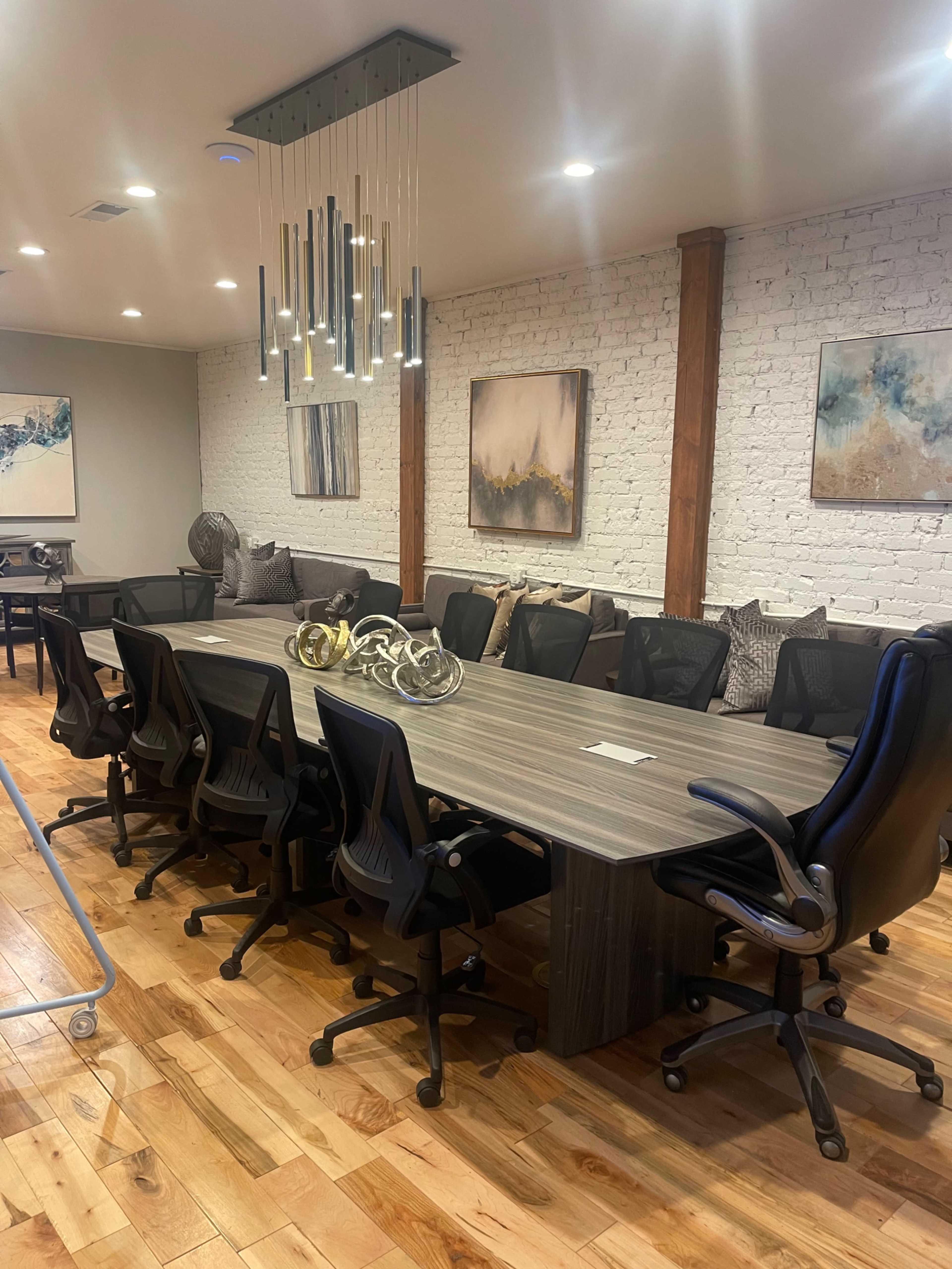 A long conference table surrounded by black ergonomic chairs is set in a well-lit room with a wooden floor and white brick walls, adorned with abstract artwork.