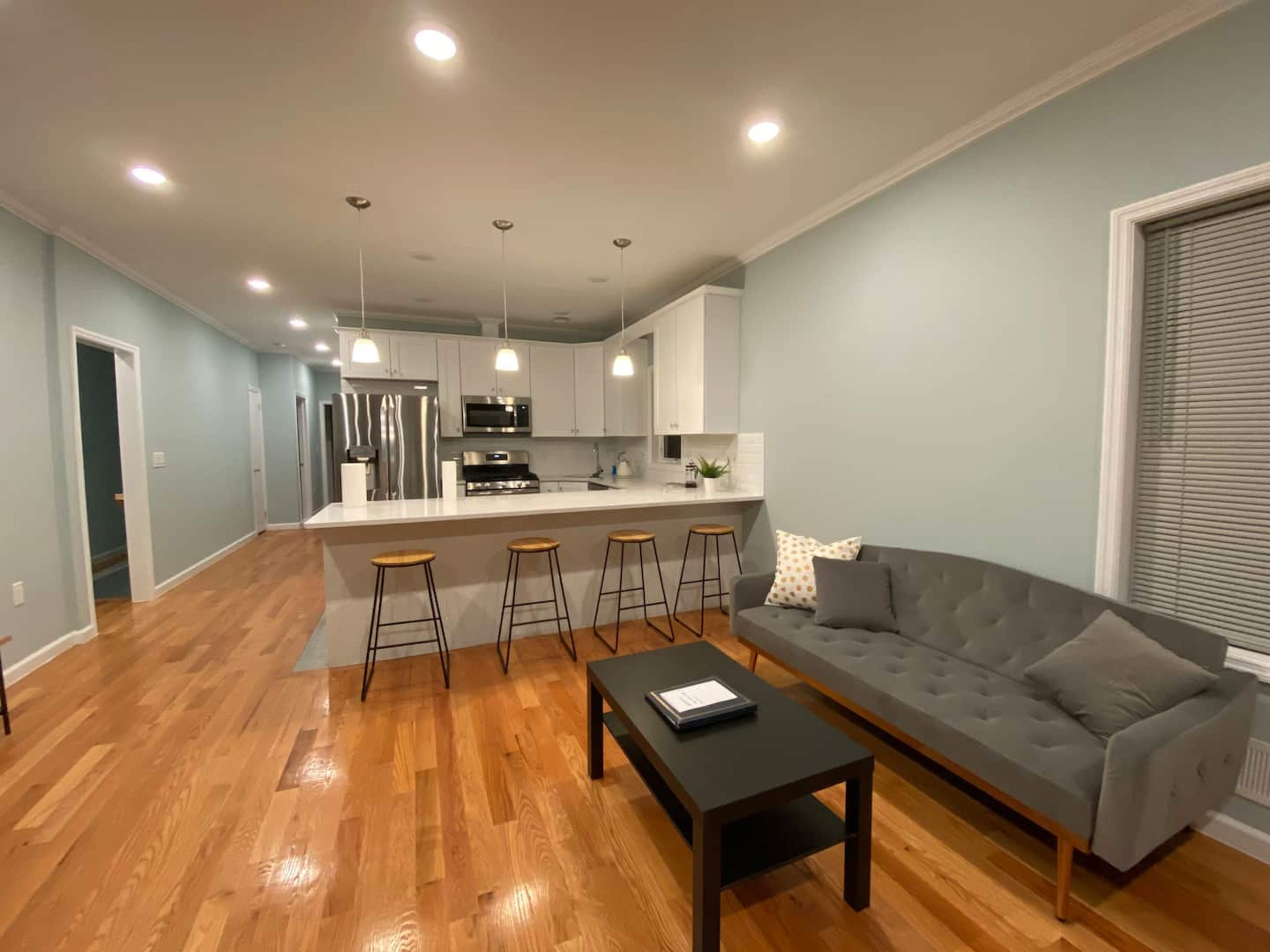 A modern living space with a gray sofa, a black coffee table, and a kitchen area featuring white cabinetry and bar stools.
