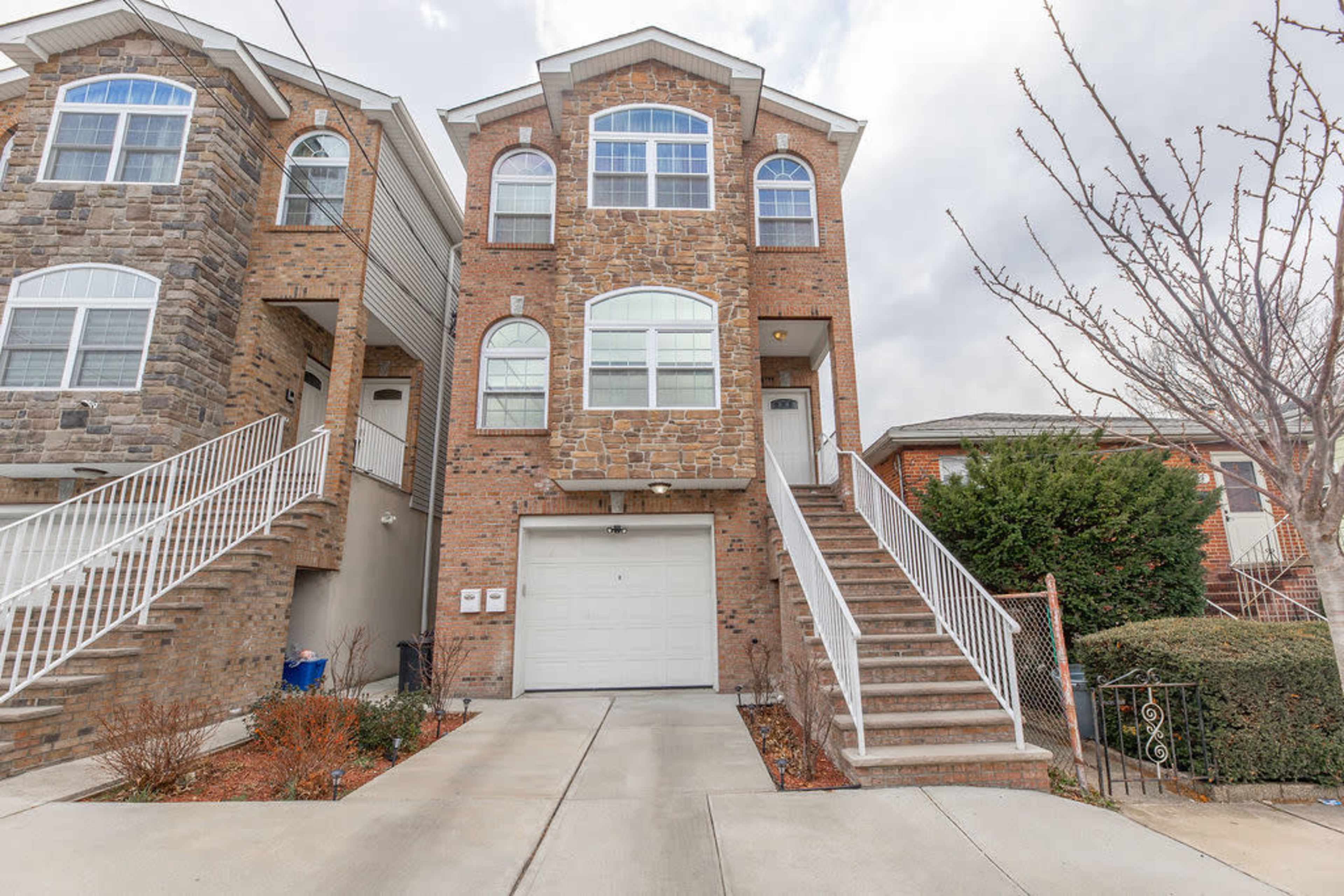 A two-story brick townhouse with a garage and front steps, bordered by another similar structure and landscaped greenery.