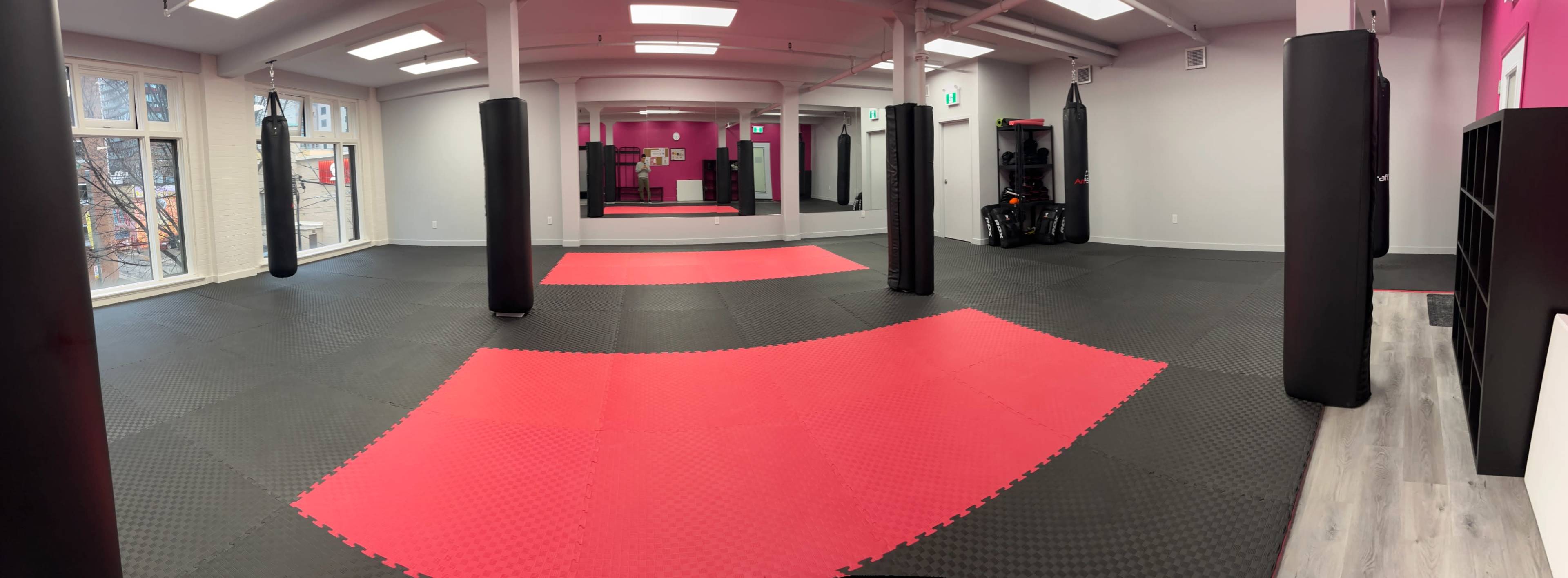 The image shows a spacious training area with black and red martial arts mats, equipped with freestanding punching bags and mirrors along the walls.
