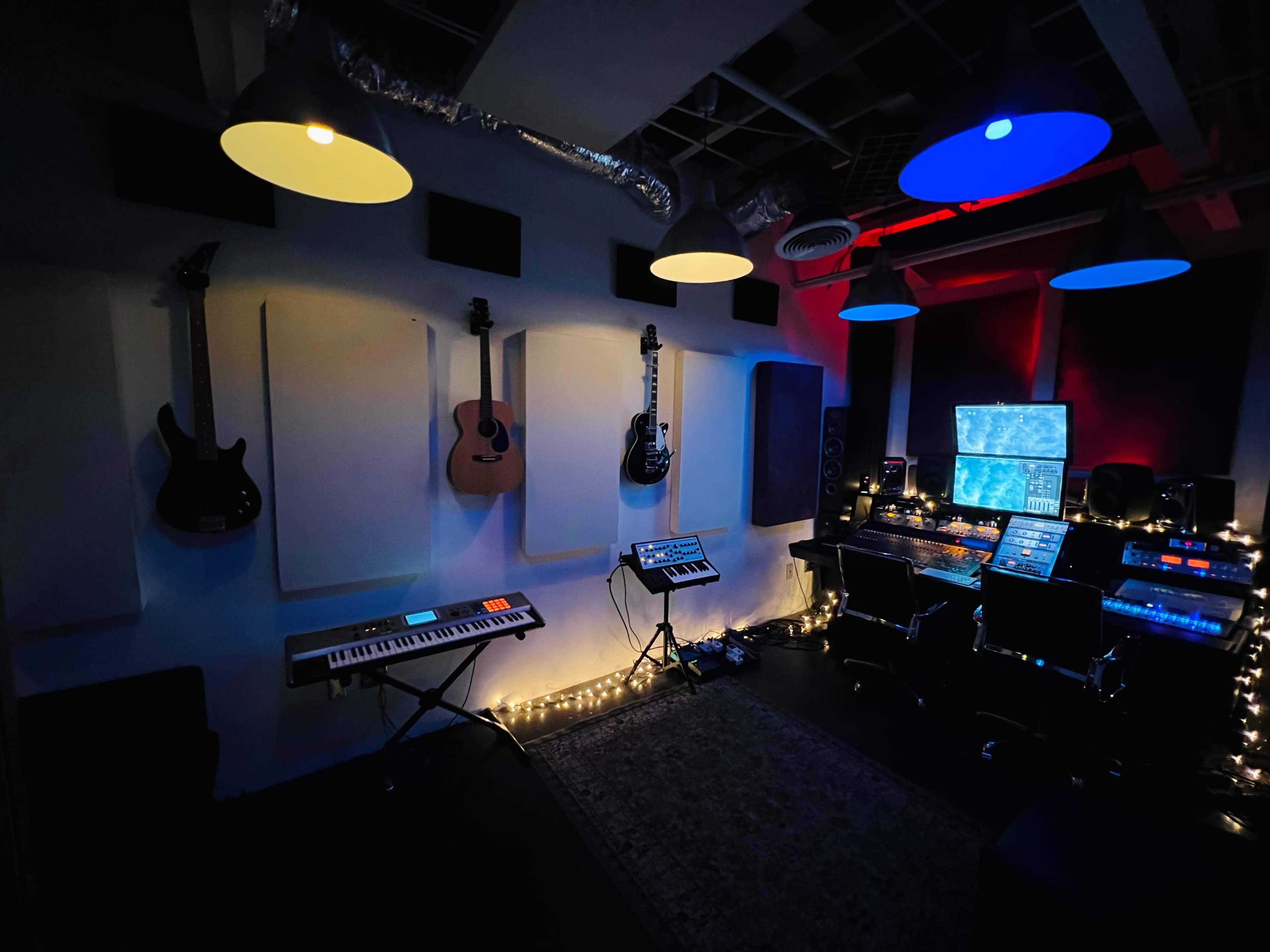 A music studio is illuminated by overhead lights, featuring guitars mounted on the walls and a workstation with various electronic equipment and a keyboard.