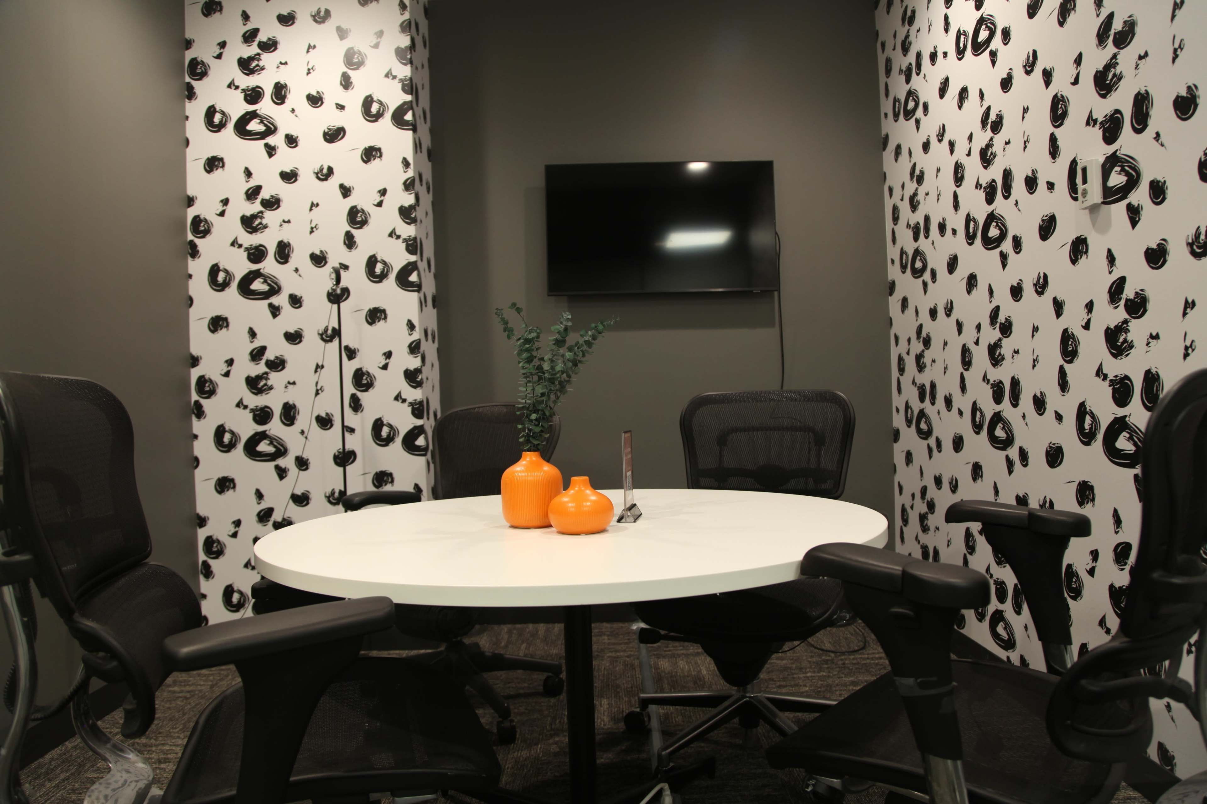 A conference room features a round white table surrounded by black office chairs, with patterned walls and a television mounted on one side.