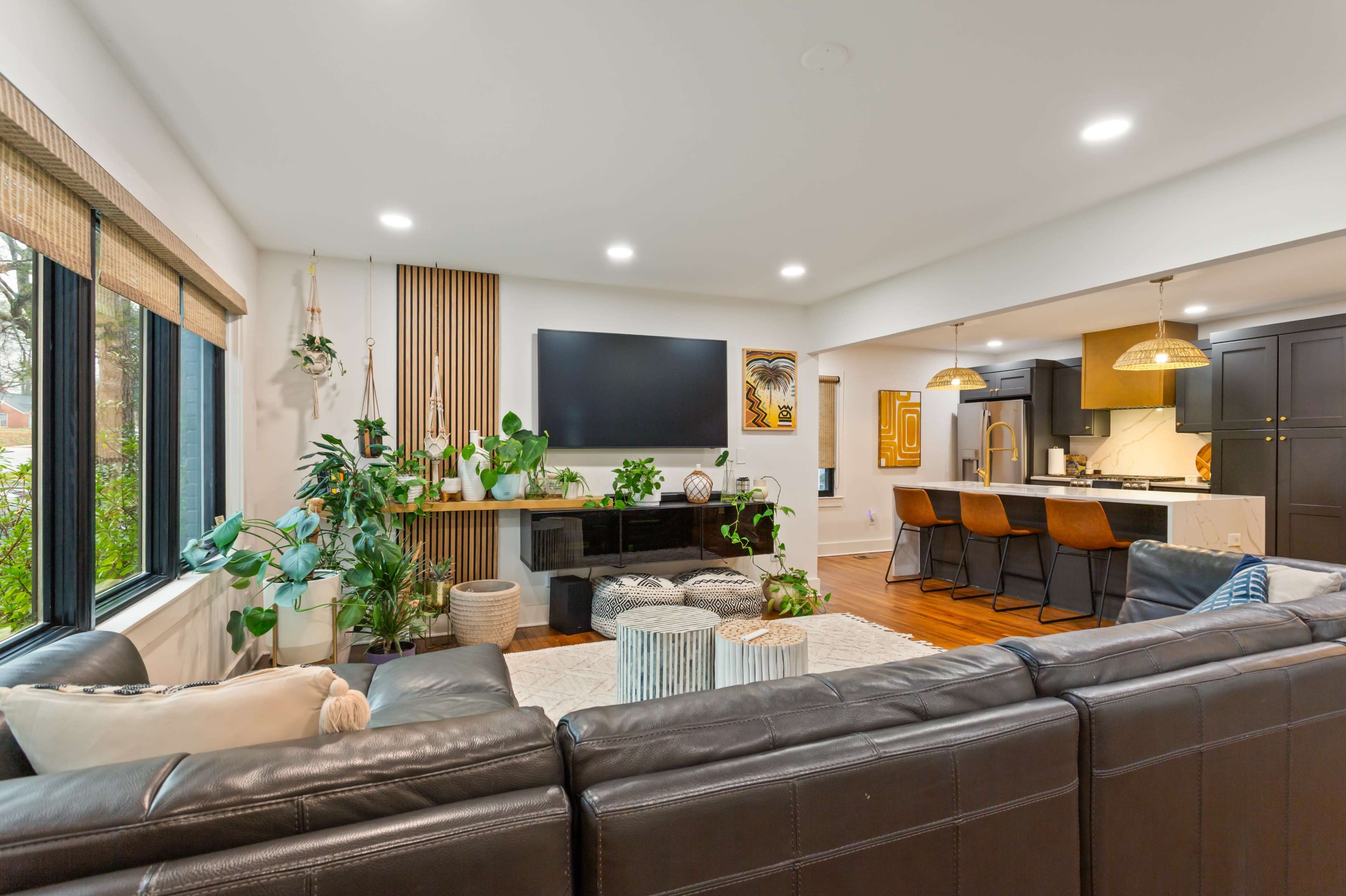 A modern living room is furnished with a dark leather sofa, potted plants, and a wall-mounted television, connecting to an open kitchen area with barstool seating.