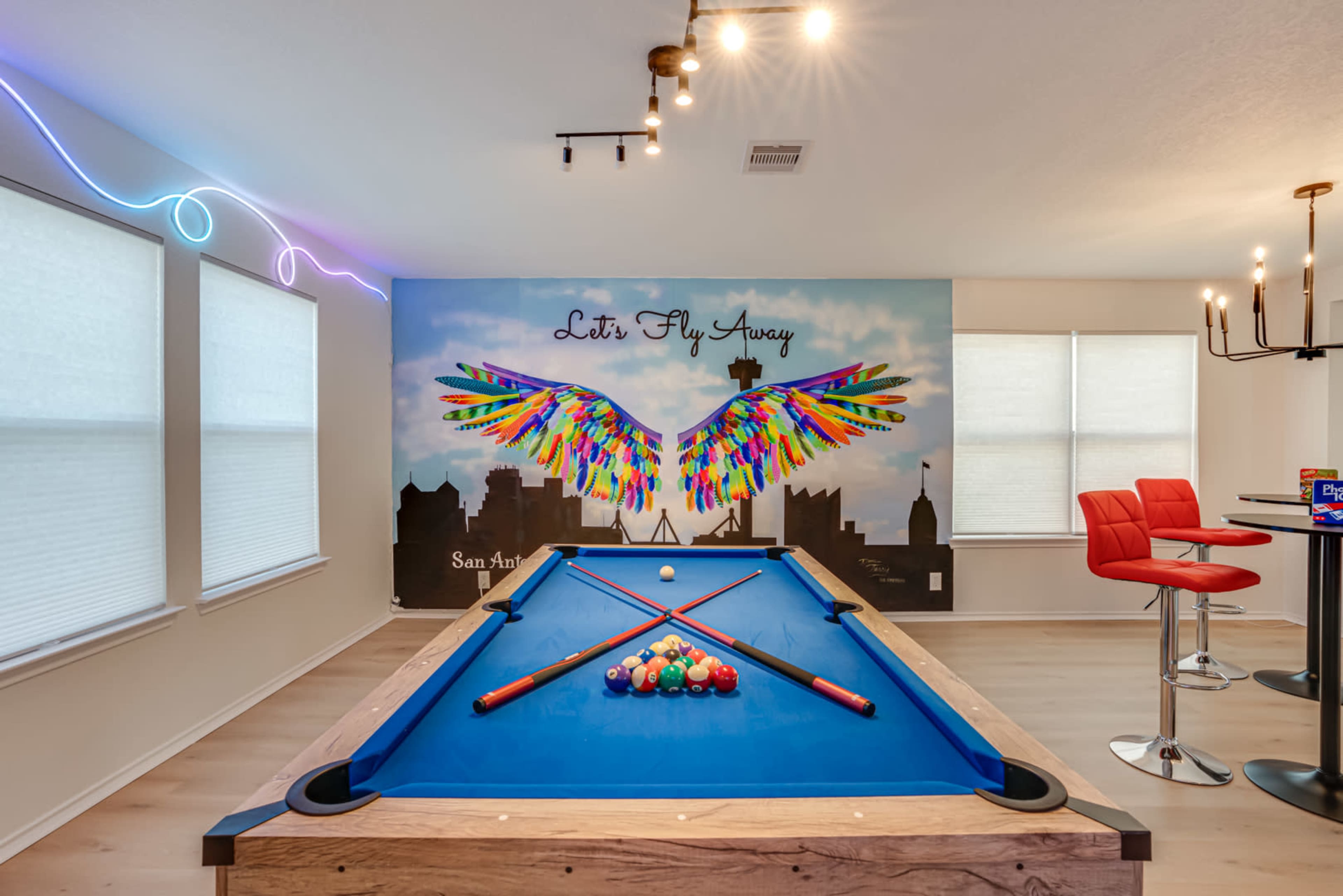 A pool table with colorful murals featuring a winged design is set against a well-lit wall with large windows and modern bar stools.