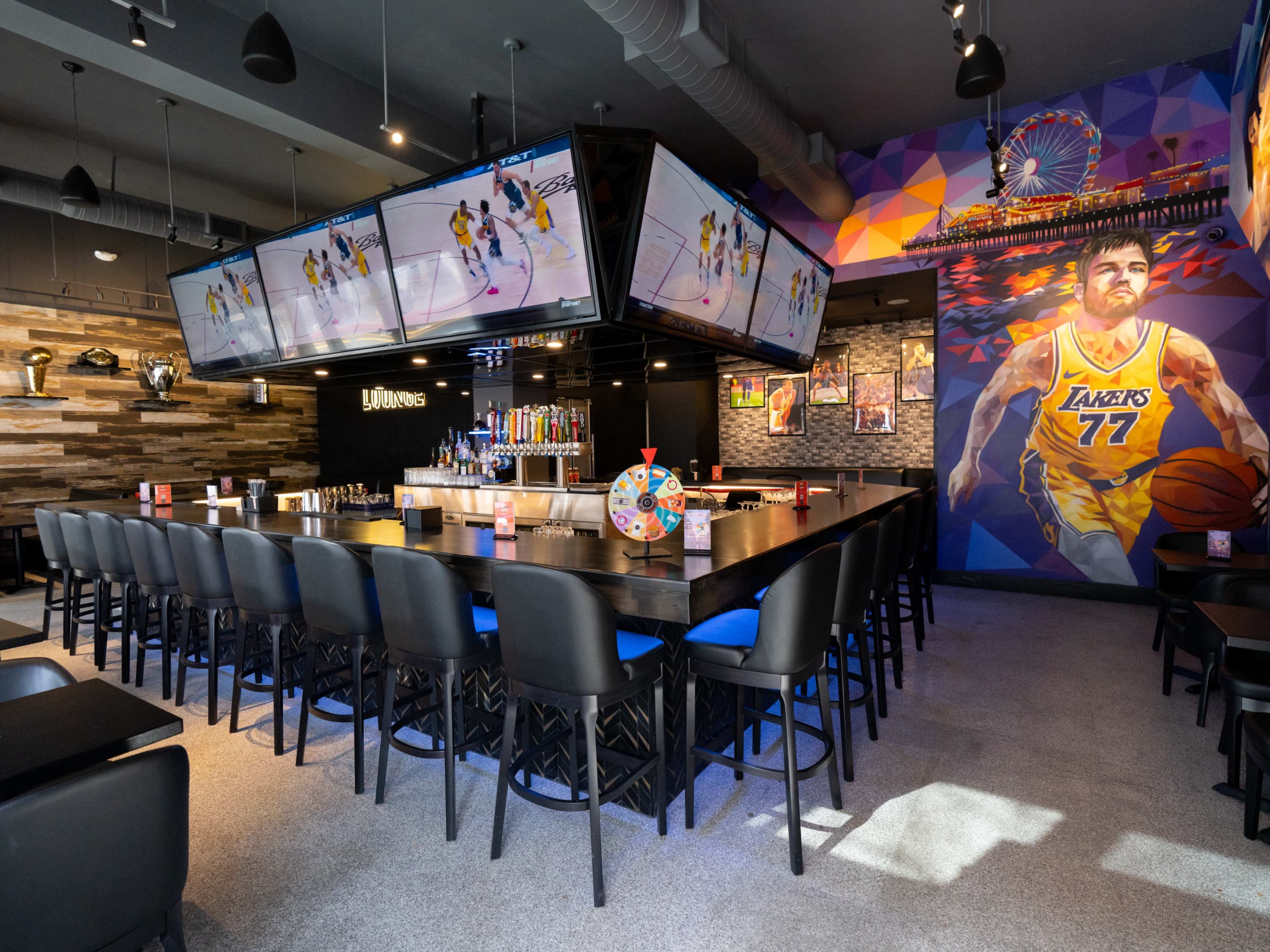 The image shows a modern sports bar with multiple large screens displaying a basketball game, a long bar with stools, and colorful wall art featuring a basketball theme.