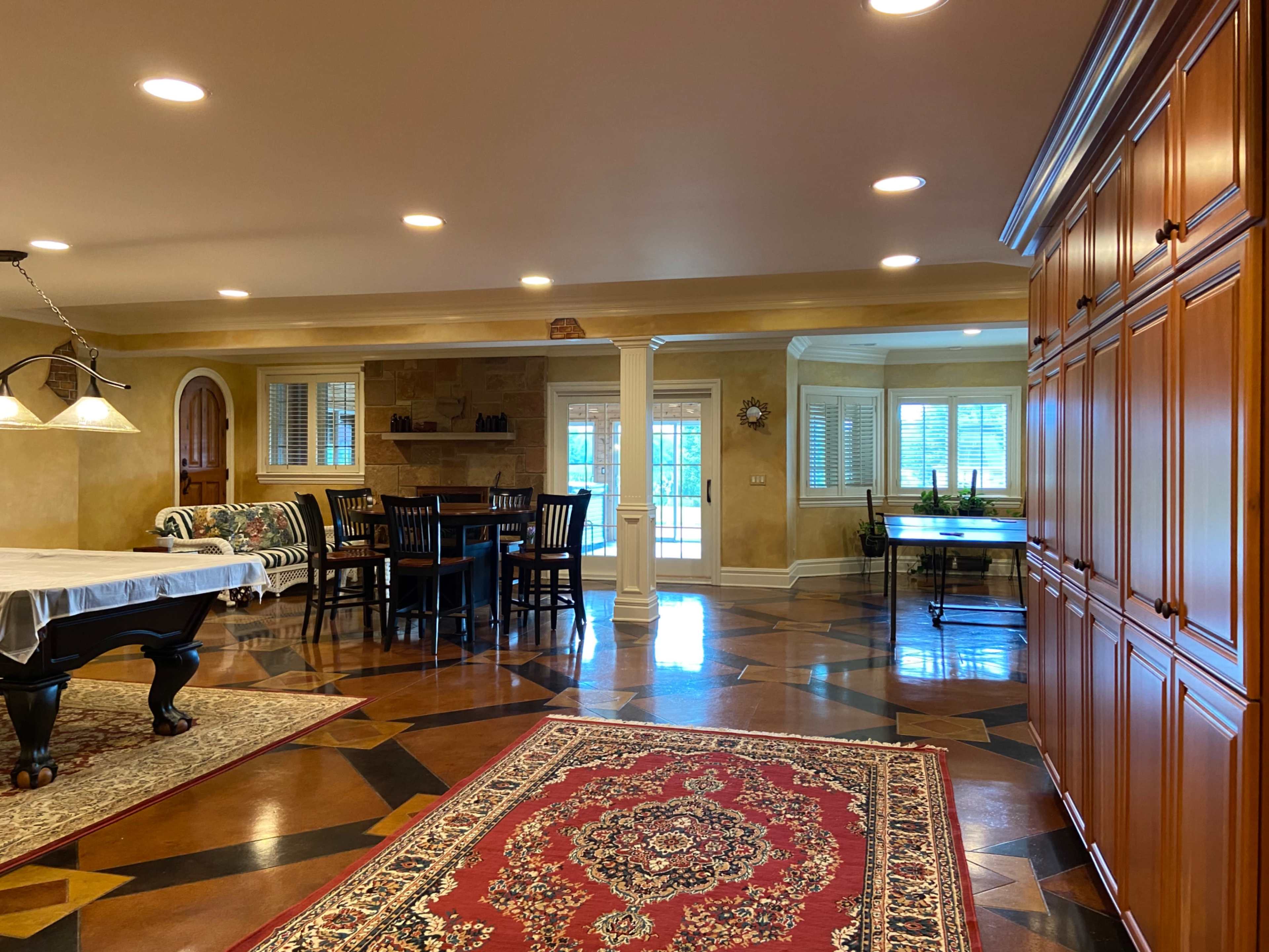 A spacious dining area featuring a wooden dining table surrounded by chairs, a pool table, and a cabinet against the wall, with large windows allowing natural light.