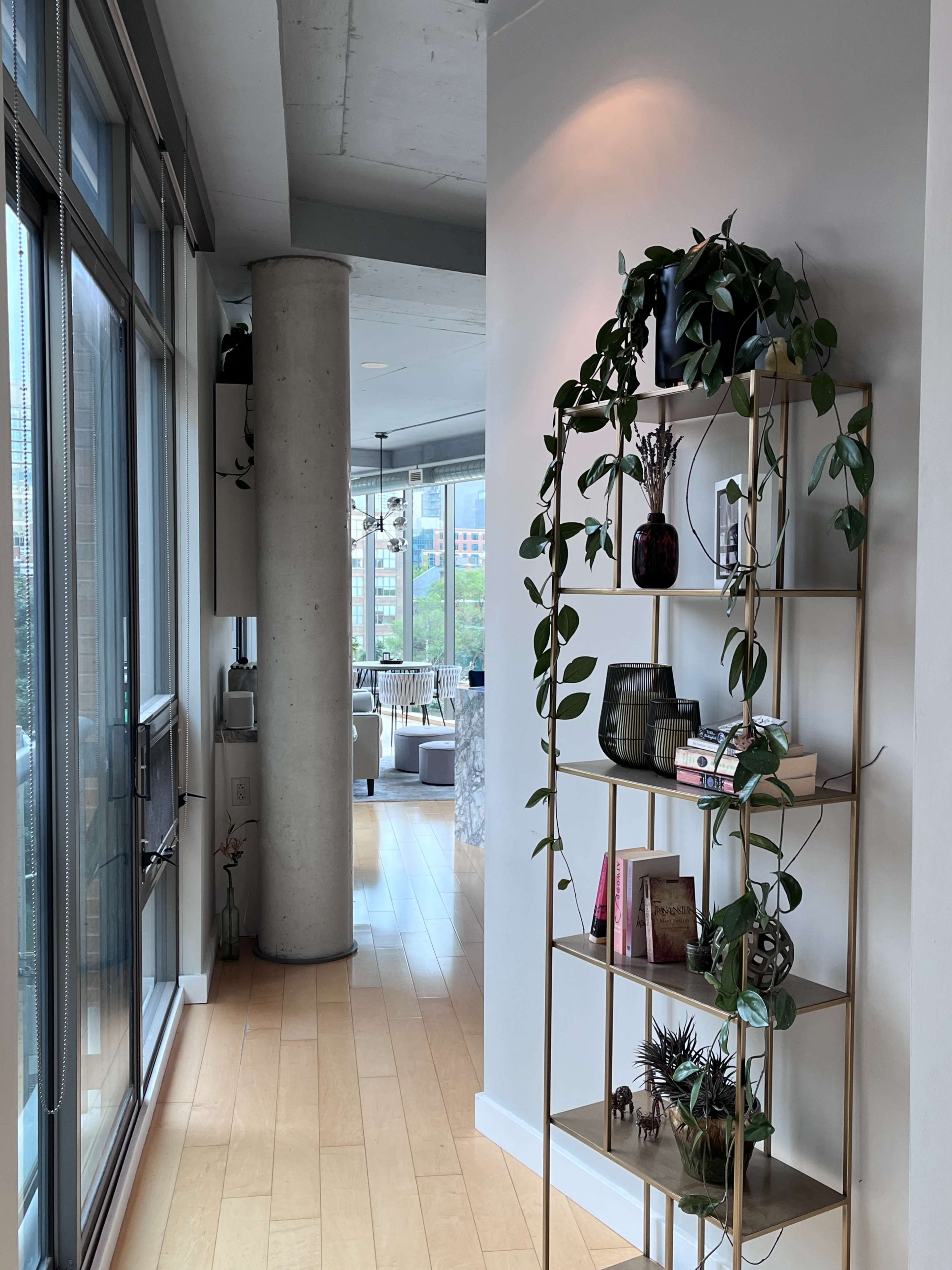 The image shows a minimalist hallway with a gold bookshelf displaying plants and books, leading to a bright living area with large windows.
