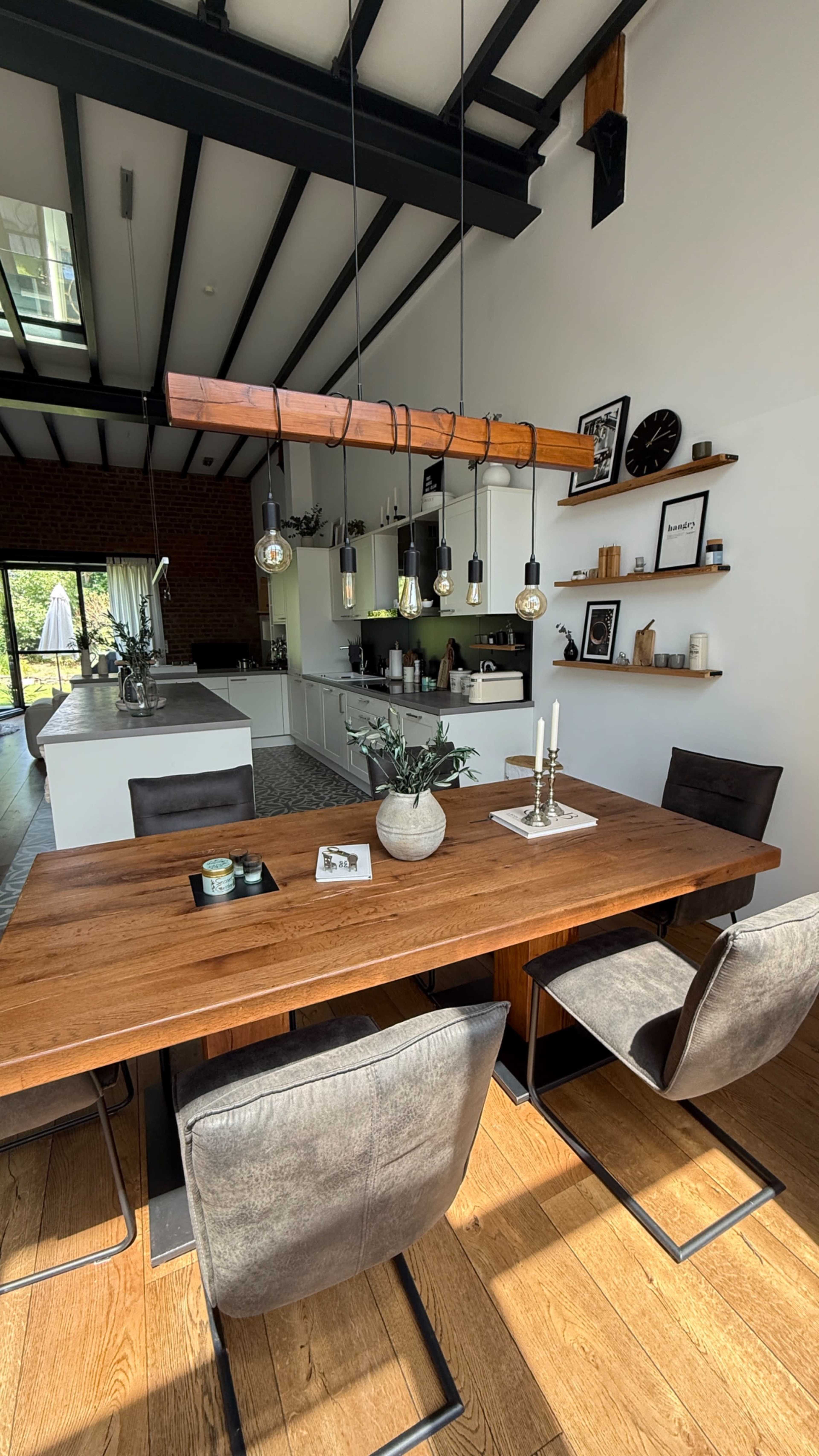 A wooden dining table with four chairs is centered in a bright room featuring an open kitchen and shelves with decor items.