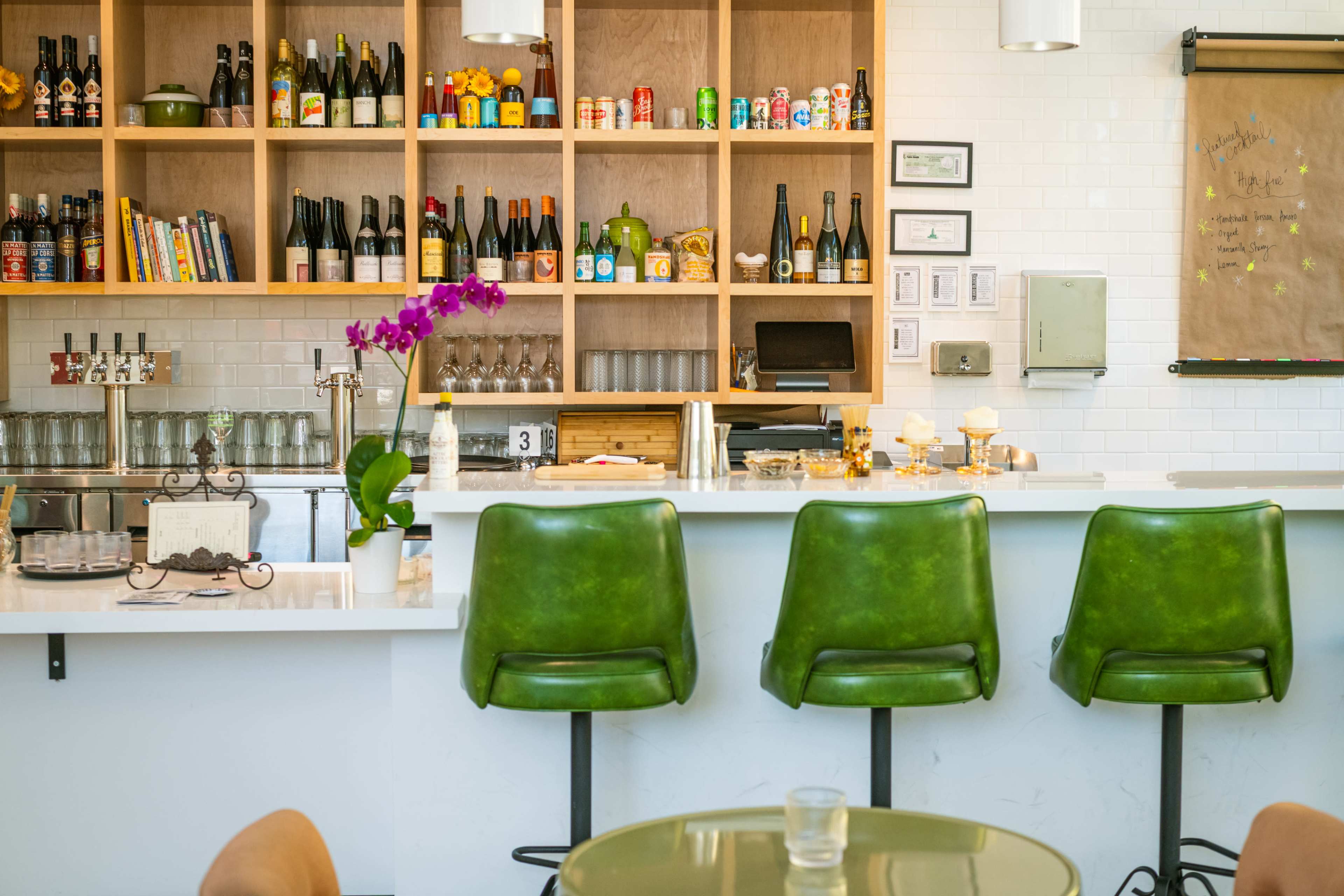 A modern bar area features three green stools in front of a white countertop, with shelves filled with various bottles and canned beverages behind it.