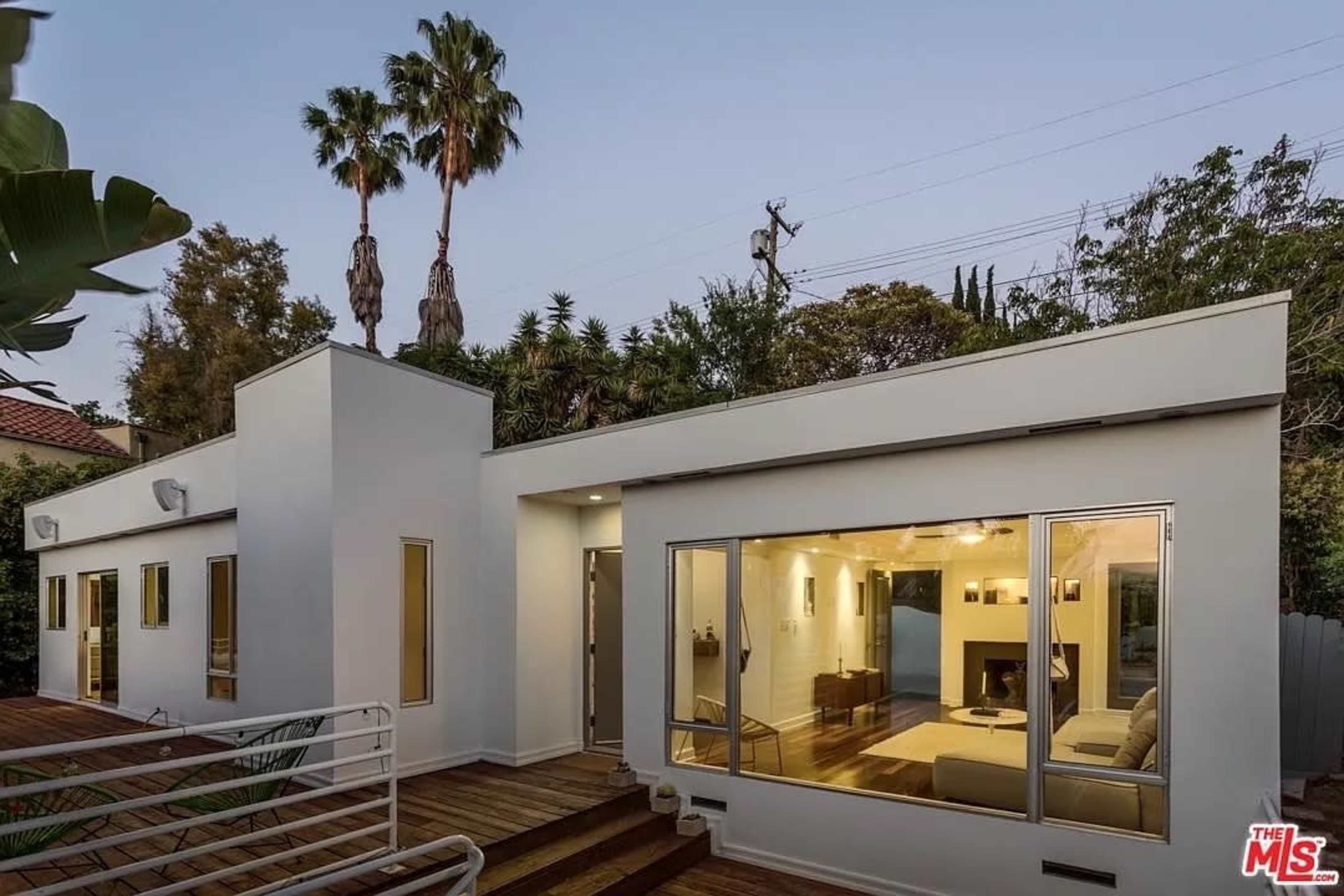 The image depicts a modern single-story house with large windows and a wooden deck, surrounded by palm trees and greenery.