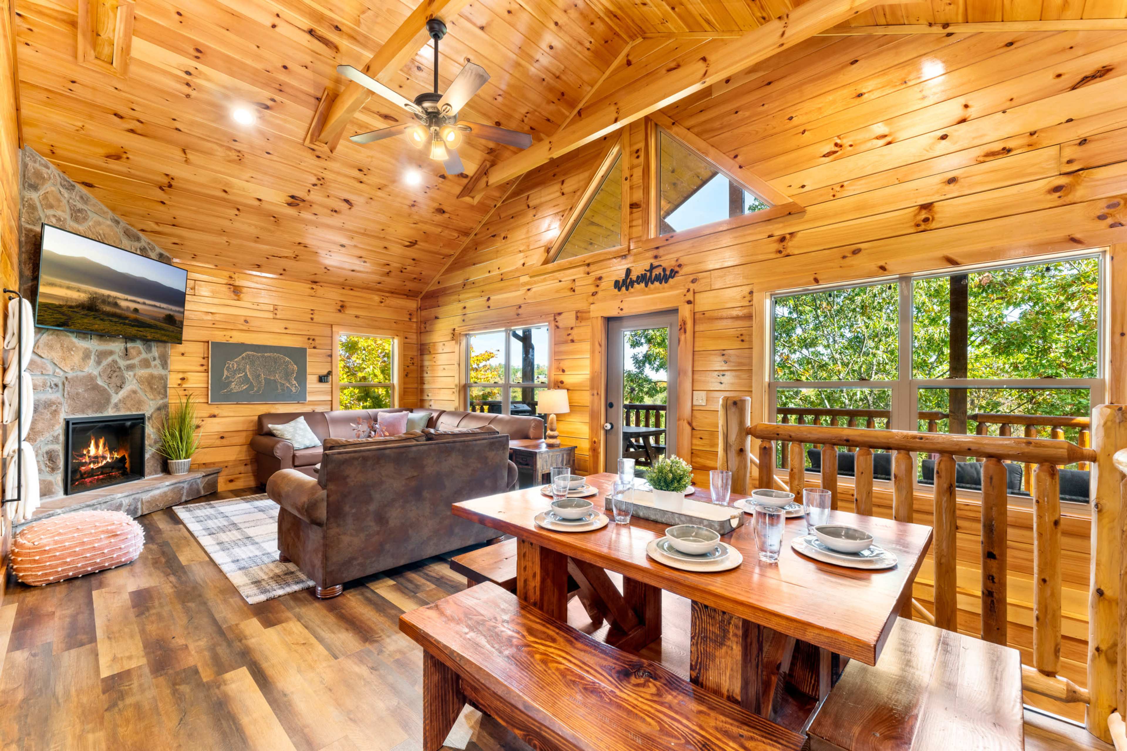 A cozy wooden cabin features a living area with a stone fireplace, a dining table, and large windows showcasing greenery outside.