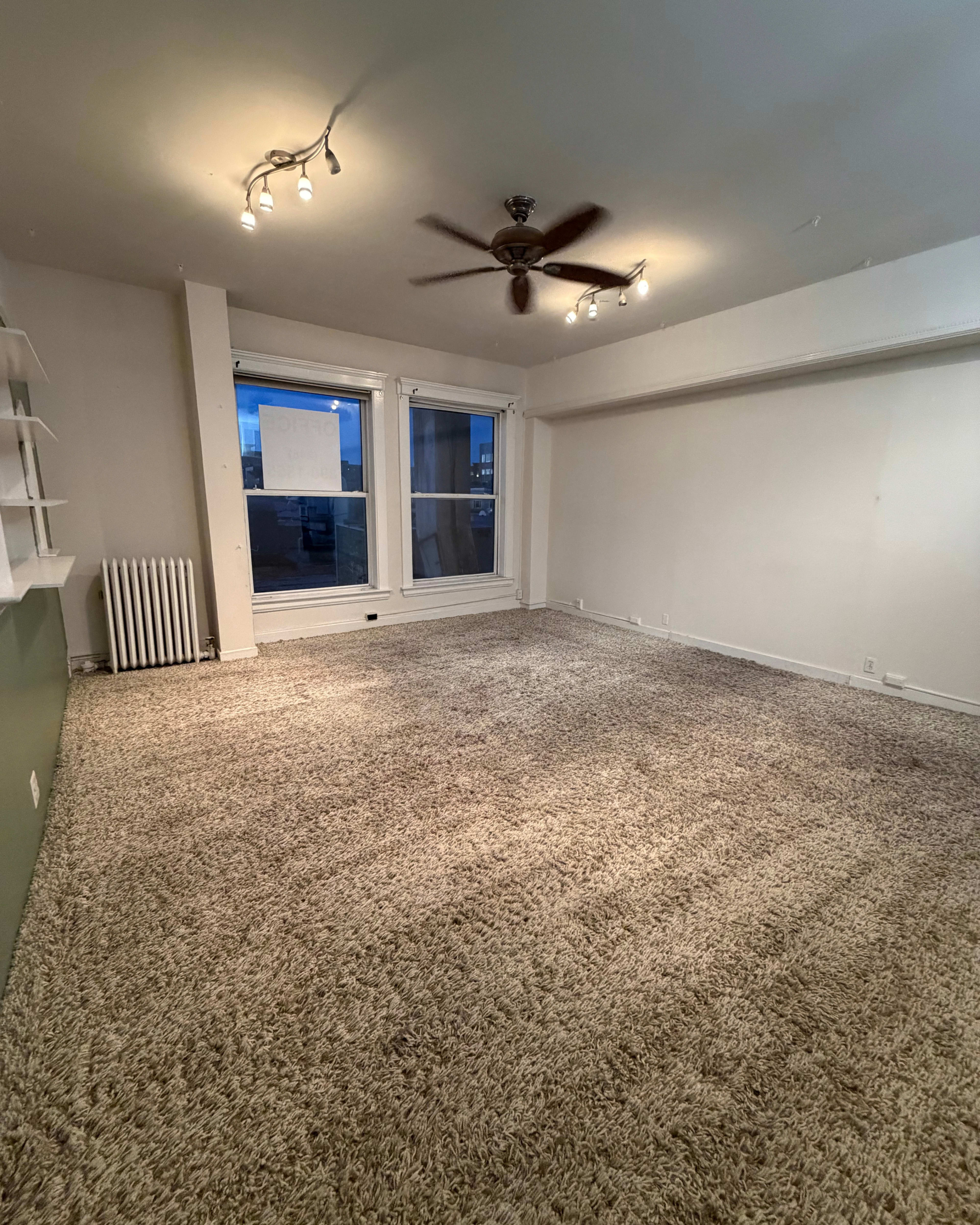 The image shows an empty room with light-colored carpet, a ceiling fan, and large windows letting in natural light.