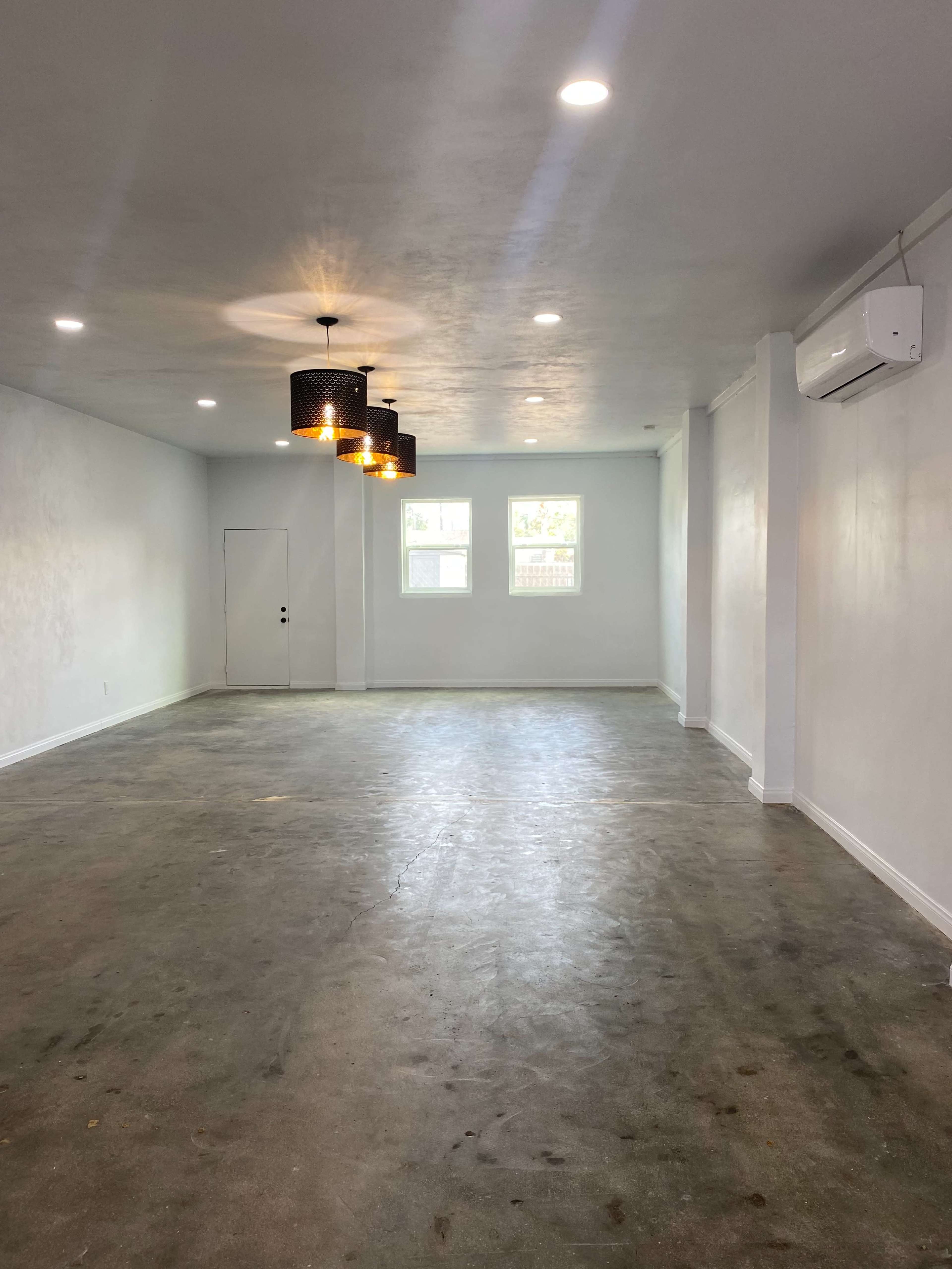 The image shows an empty room with a concrete floor, two windows, and a ceiling featuring hanging light fixtures.