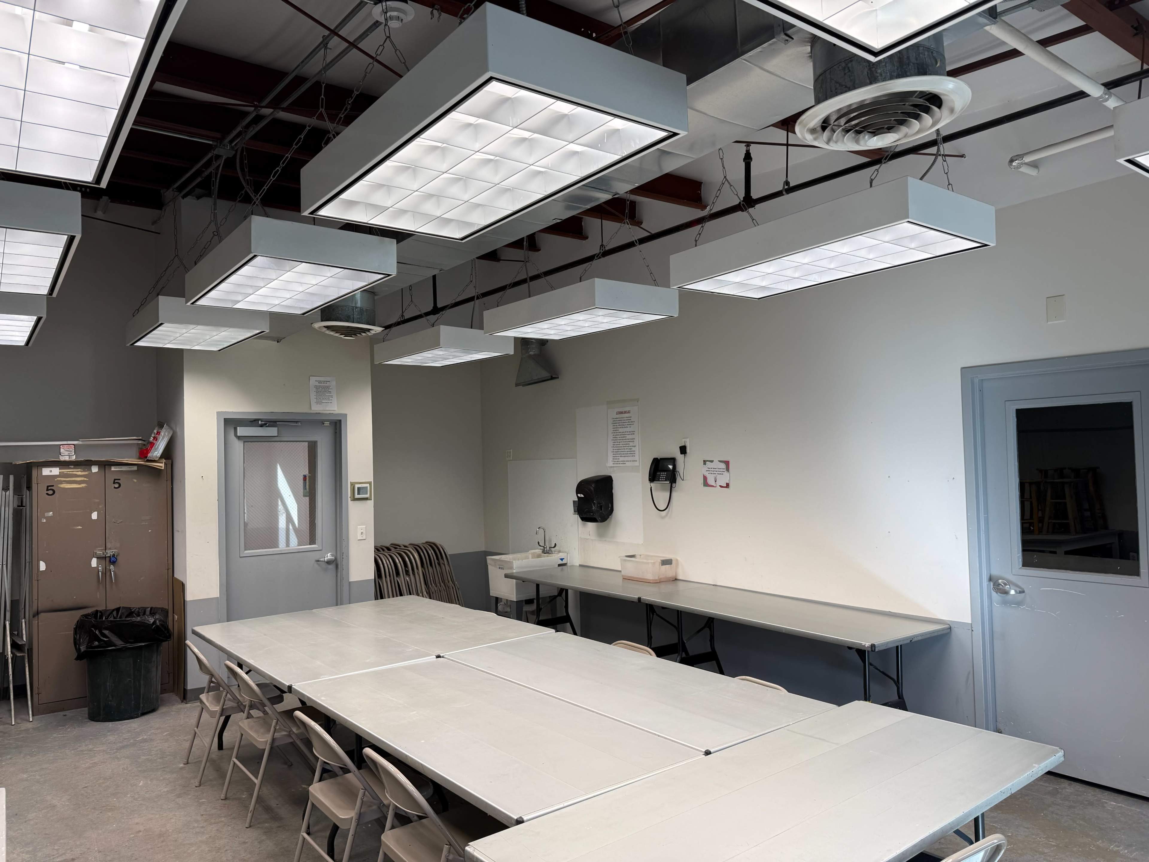 The image shows a well-lit classroom with several overhead light fixtures, a long table with chairs, and a door leading to another room.