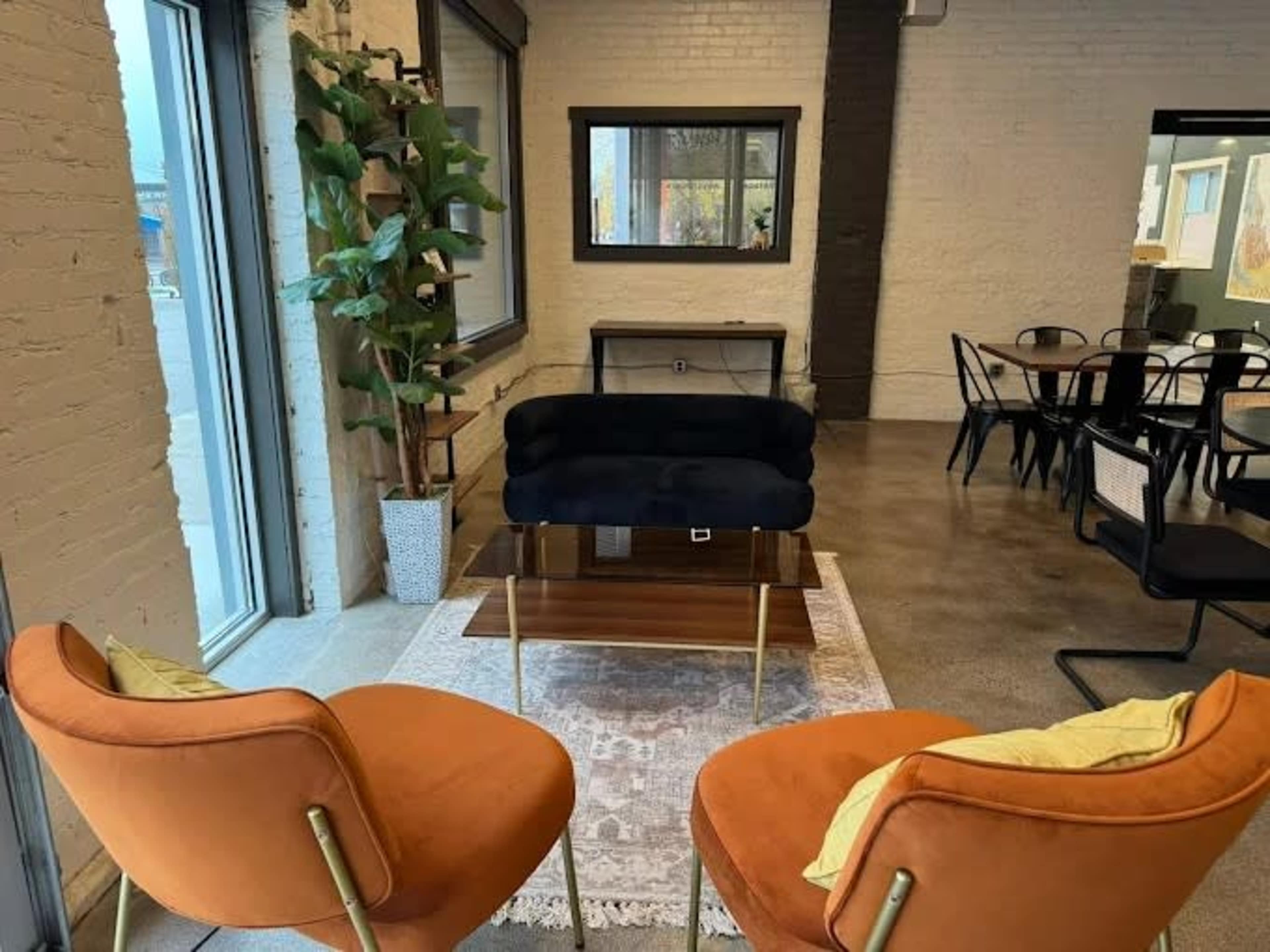 The image shows a modern seating area with two orange chairs facing a black couch, a coffee table in the center, and large windows letting in natural light.
